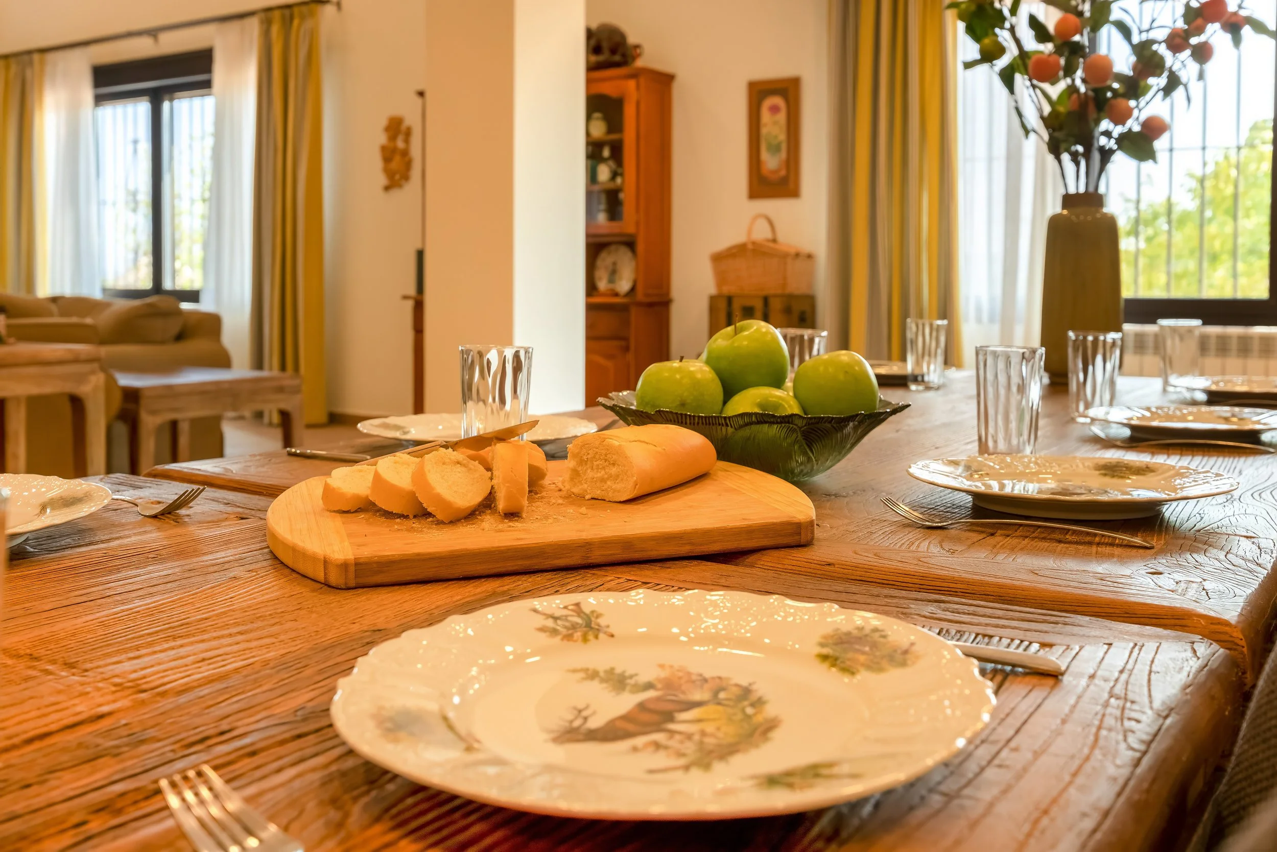 Een eettafel met borden, glazen en bestek, een houten snijplank met brood en kaas, een kom met groene appels, en een hoge vaas met een grote plant met oranje vruchten, in een gezellige kamer met grote ramen, gordijnen en houten meubels.