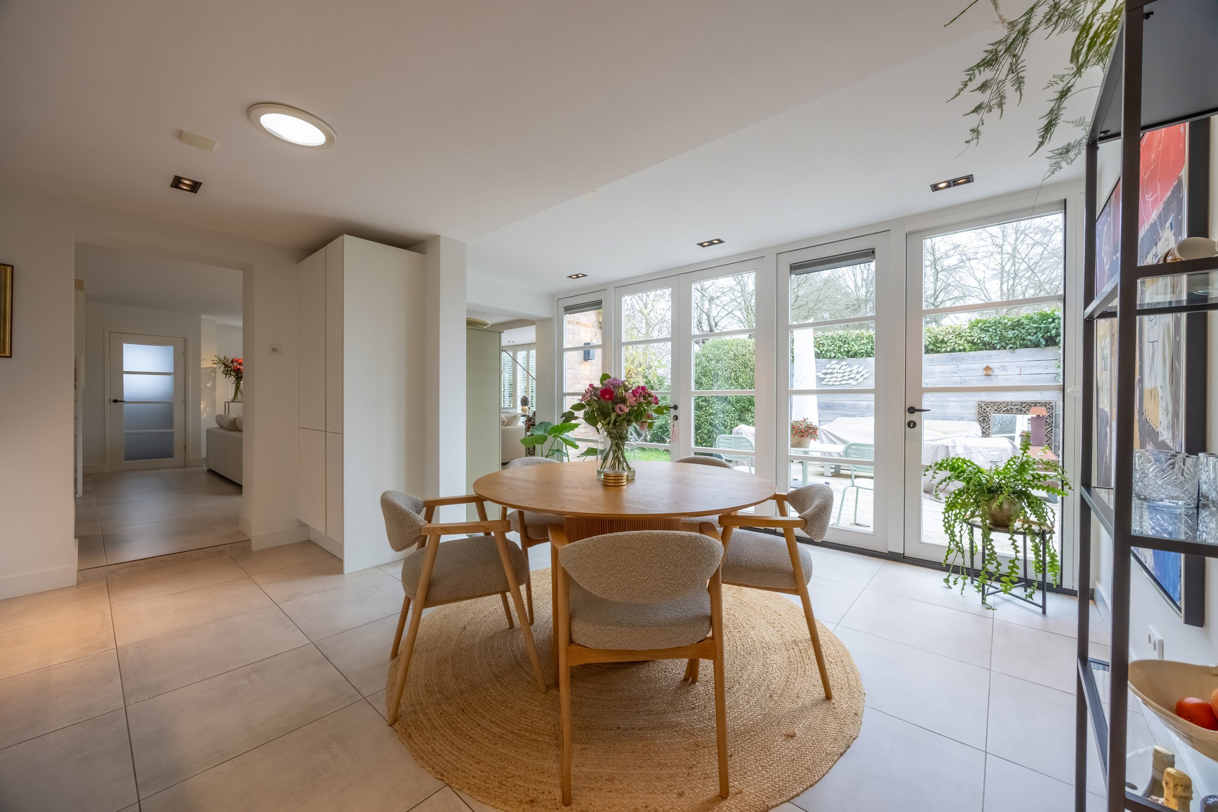 Eethoek met ronde houten tafel, zes stoffen stoelen, bloembol in vazen, grote ramen uitzicht naar tuin.