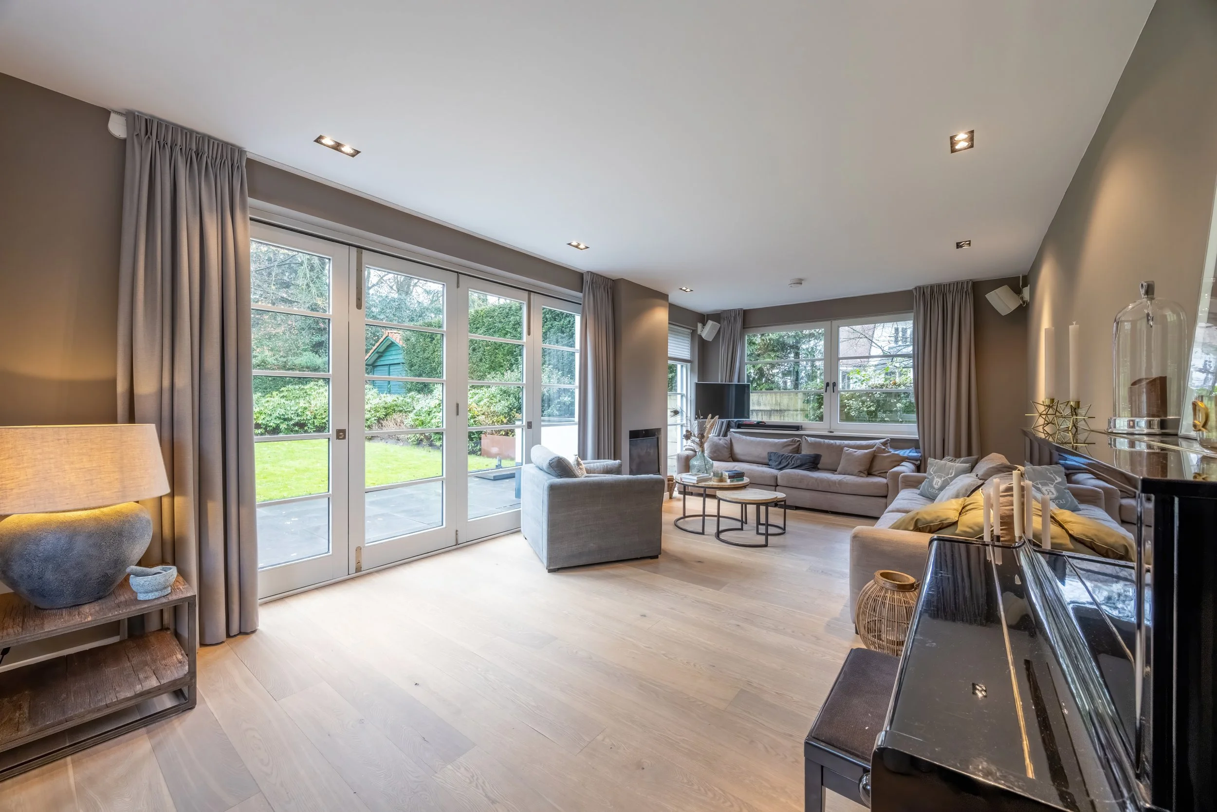 Lichte woonkamer met grote ramen en deur naar tuin, hoekbank, salontafels, open haard, piano, en decoratieve accessoires.