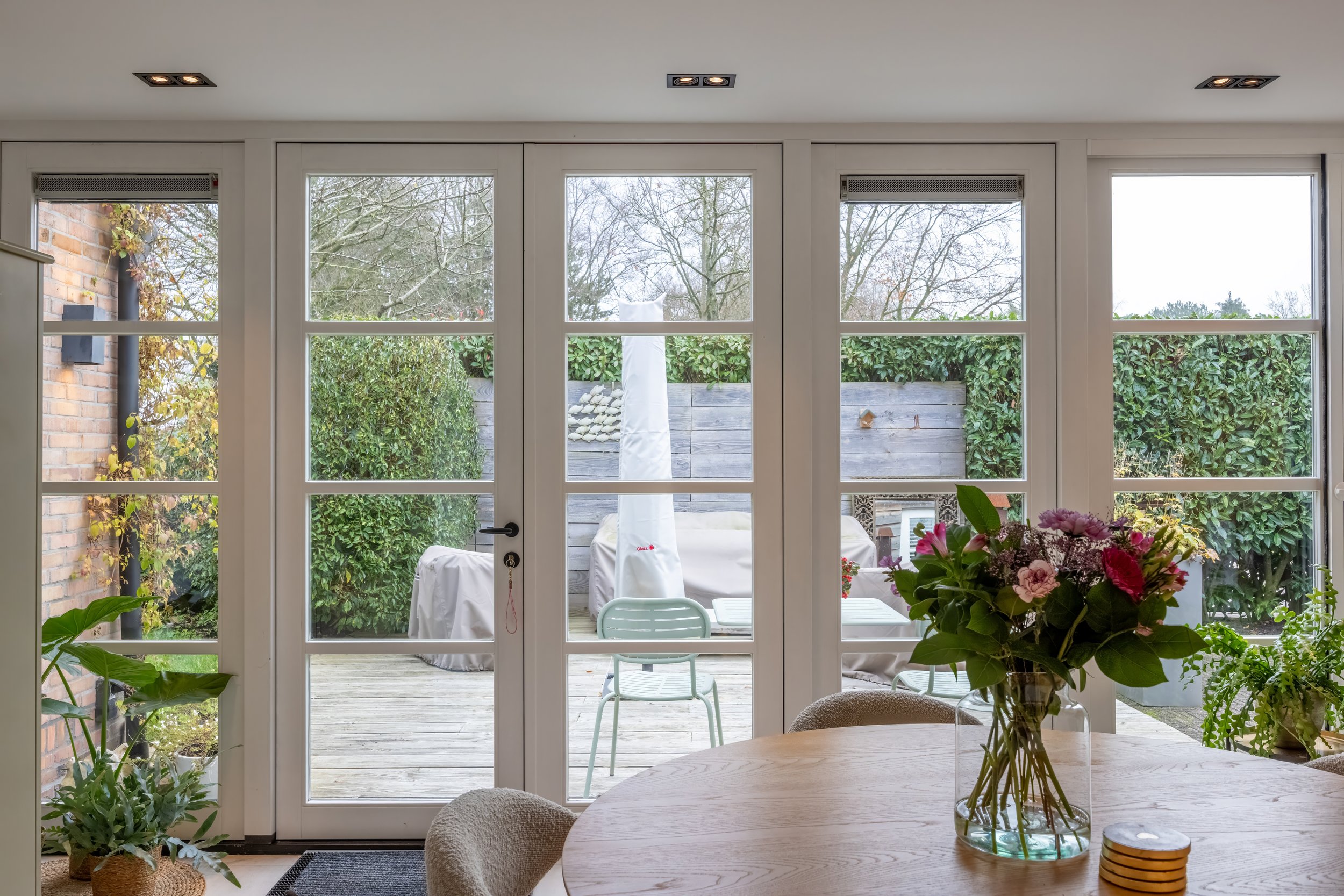 Binnenzicht op een woonkamer met grote glazen deuren die uitkijken op een terras met tuin en planten.