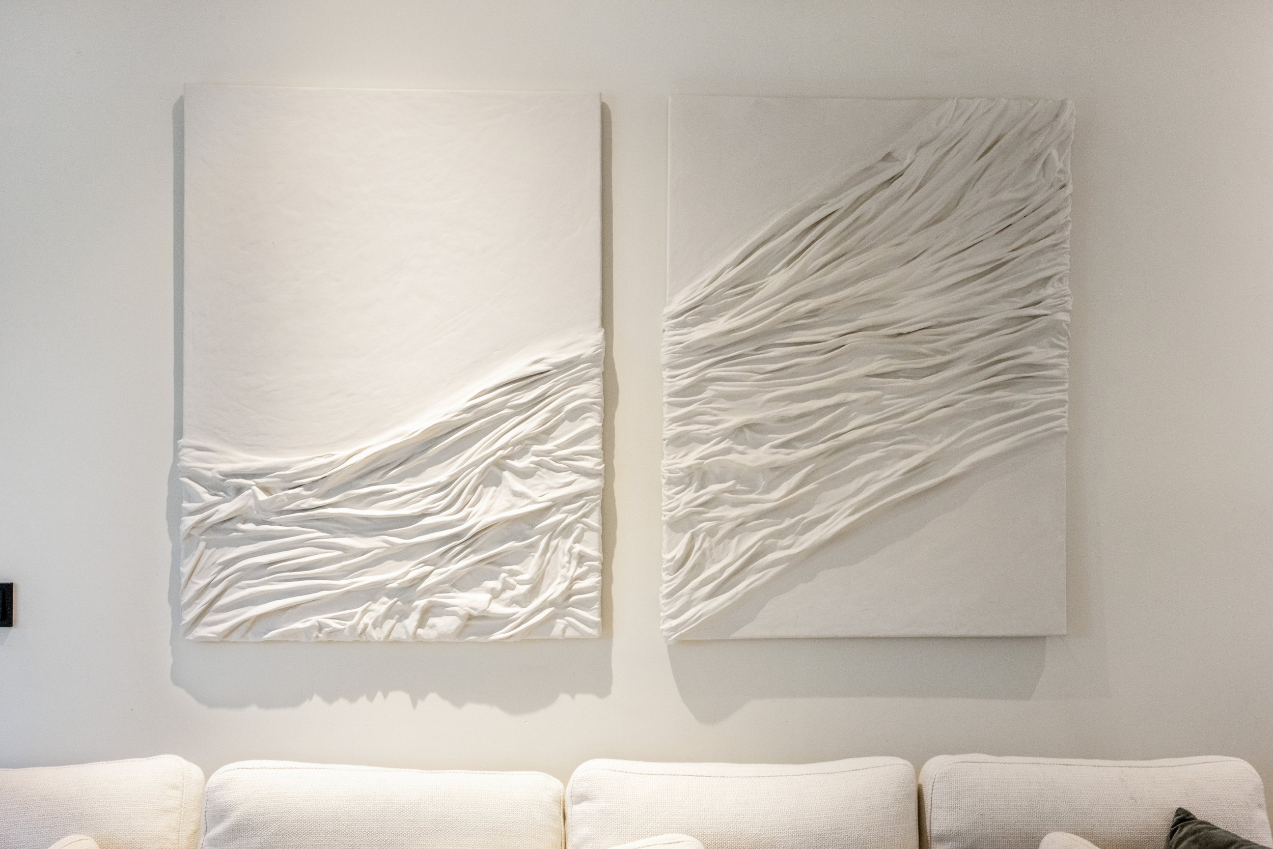 Twee witte 3D-wandkunstwerken met gerolde textuurlijnen hangen boven een witte bank.