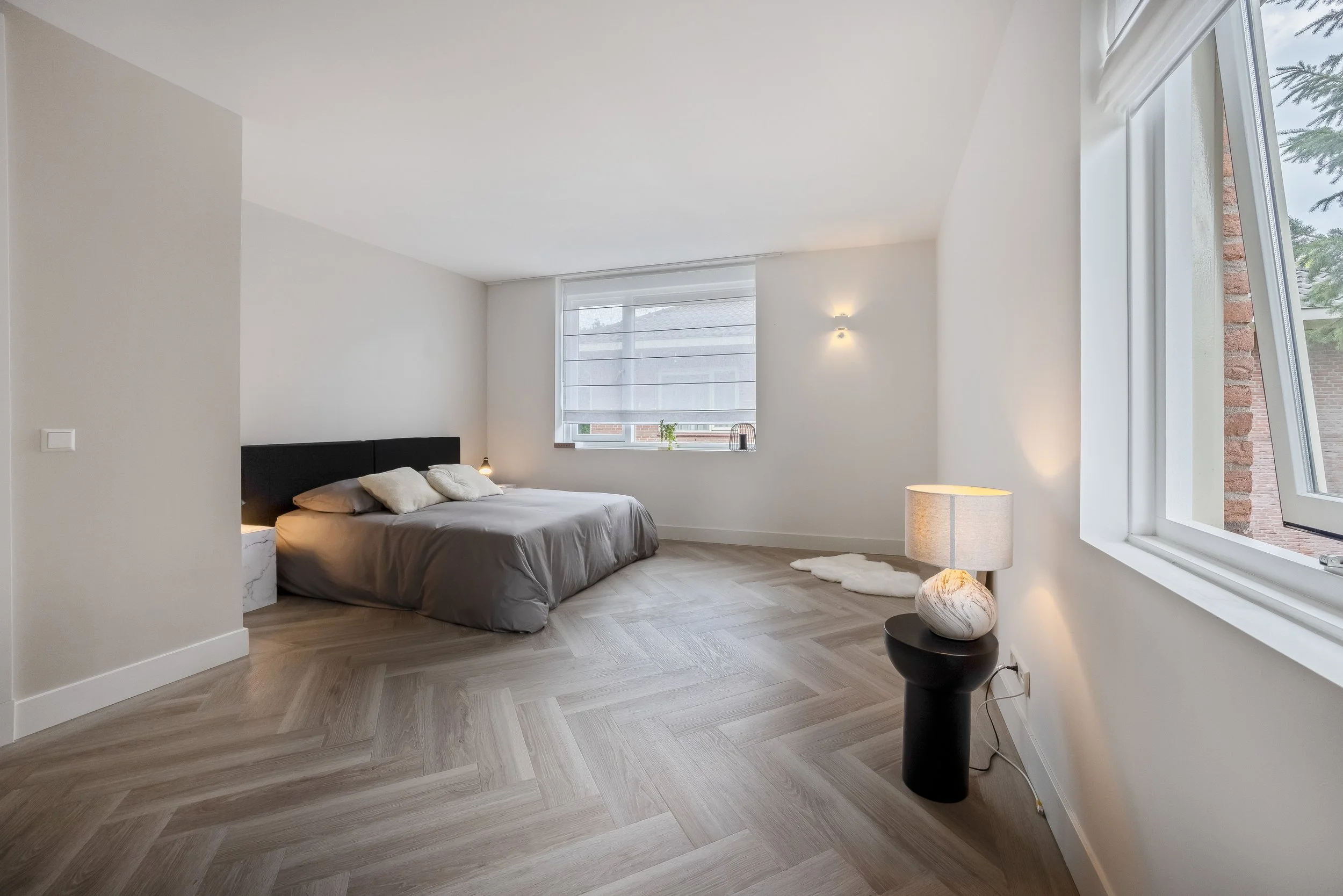 Slaapkamer met groot bed, houten vloer, grote ramen, nachtlampje en tafellamp, minimalistische inrichting