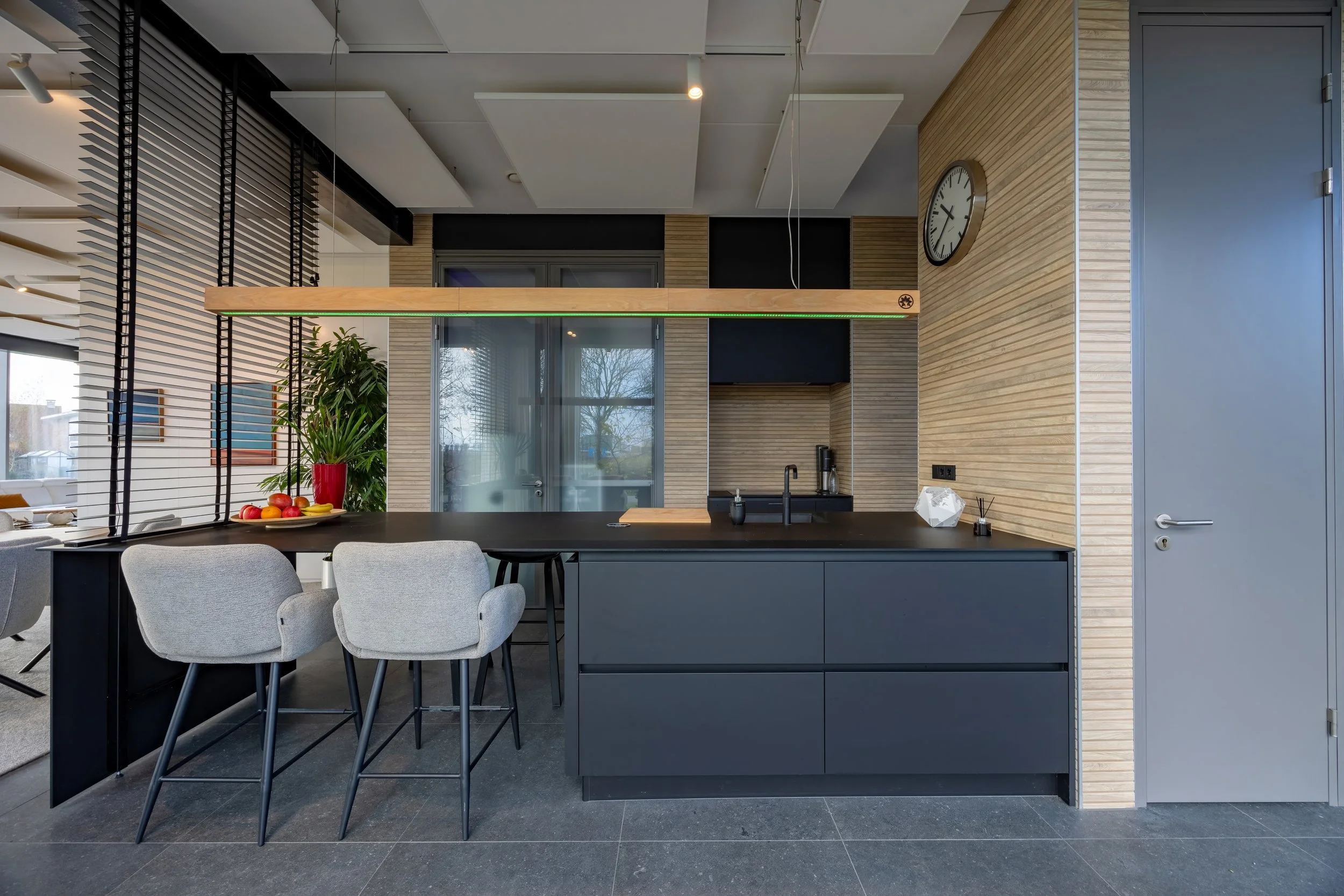 Moderne keuken met zwarte kast, houten accentwand en vier hoge stoelen, met fruit en planten op het aanrecht en een klok aan de muur.
