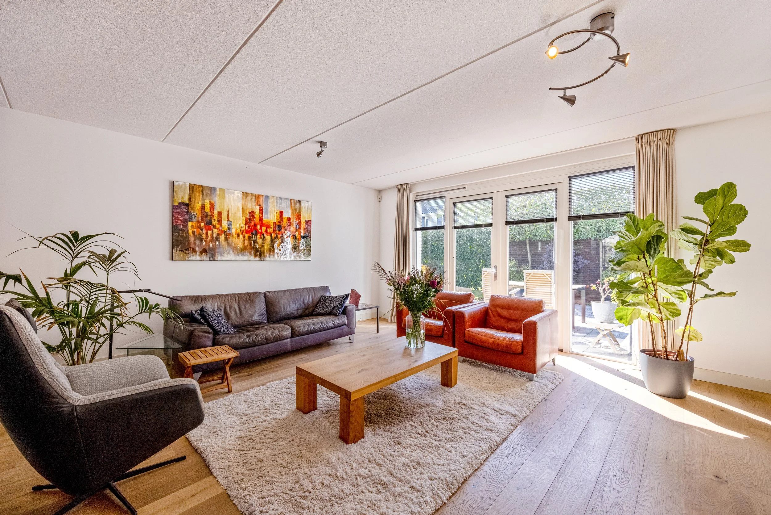 Een lichte woonkamer met grote ramen, een bruine bank, twee oranje stoelen, een lichtgrijze fauteuil, een houten salontafel met bloemstuk, een vloerkleed, planten en een moderne plafondlamp.