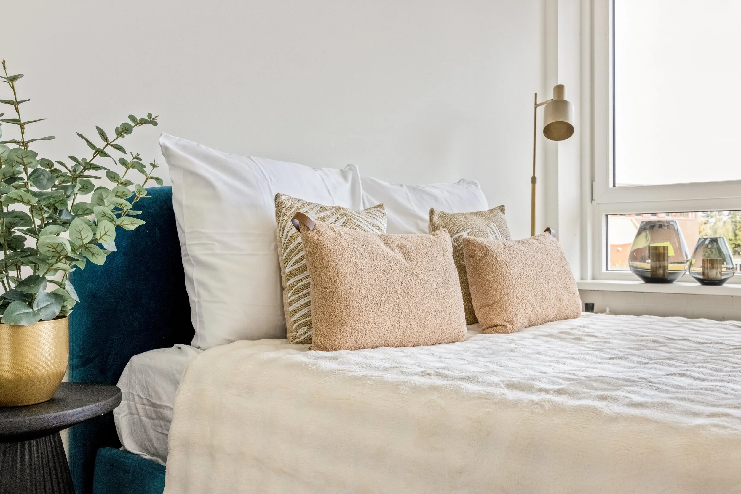 Een slaapkamer met een bed dat sierkussens heeft, een nachtkastje met een groene plant, en een raam met decoratieve vazen binnen. 