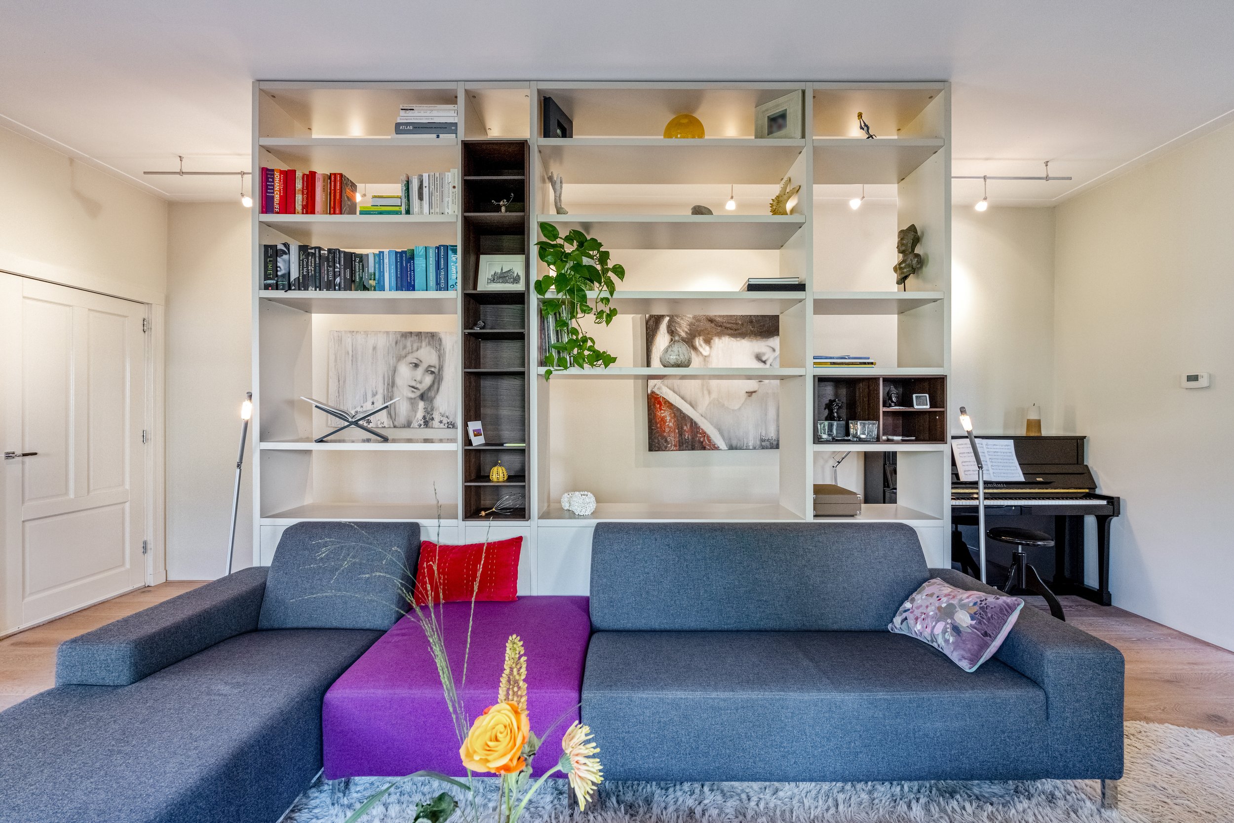 Een woonkamer met een grote boekenkast, een bank met kussens en bloemen op een tafel, een piano in de hoek, en witte muren.