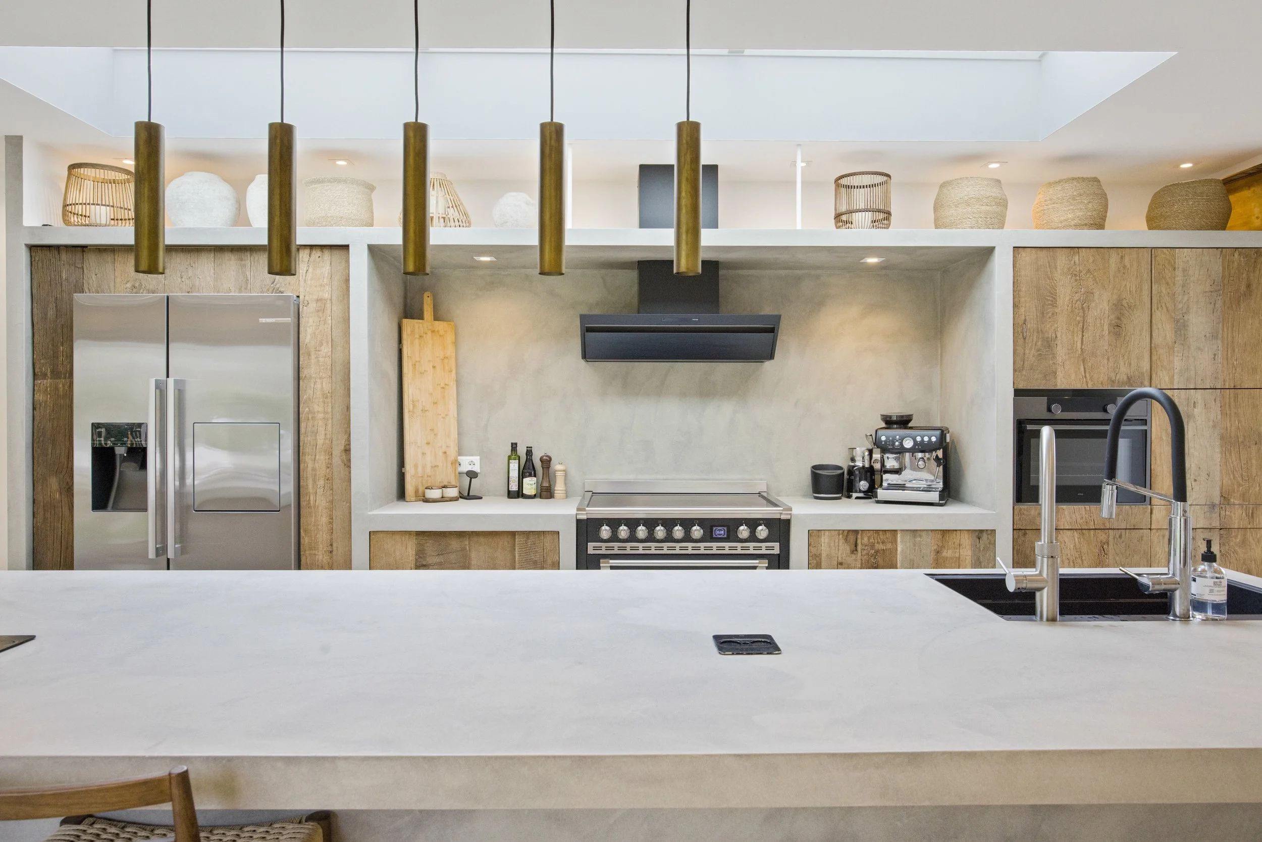 Moderne keuken met houten en betonnen elementen, keukenkastjes, een fornuis, ingebouwde oven en koelkast, en een eiland met een wastafel.