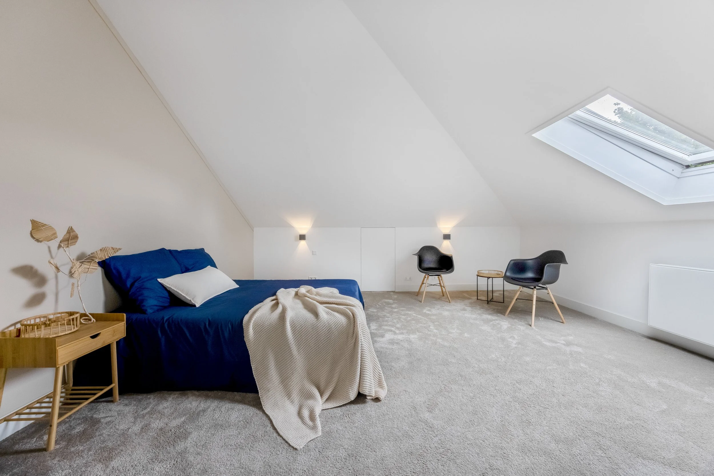 Slaapkamer met witte muren en een schuin plafond, een bed met blauwe lakens en een wit kussen, een beige plaid, een klein houten nachtkastje met een decoratief blad en bladeren, twee zwarte moderne stoelen, een klein bijzettafeltje en een groot dakra