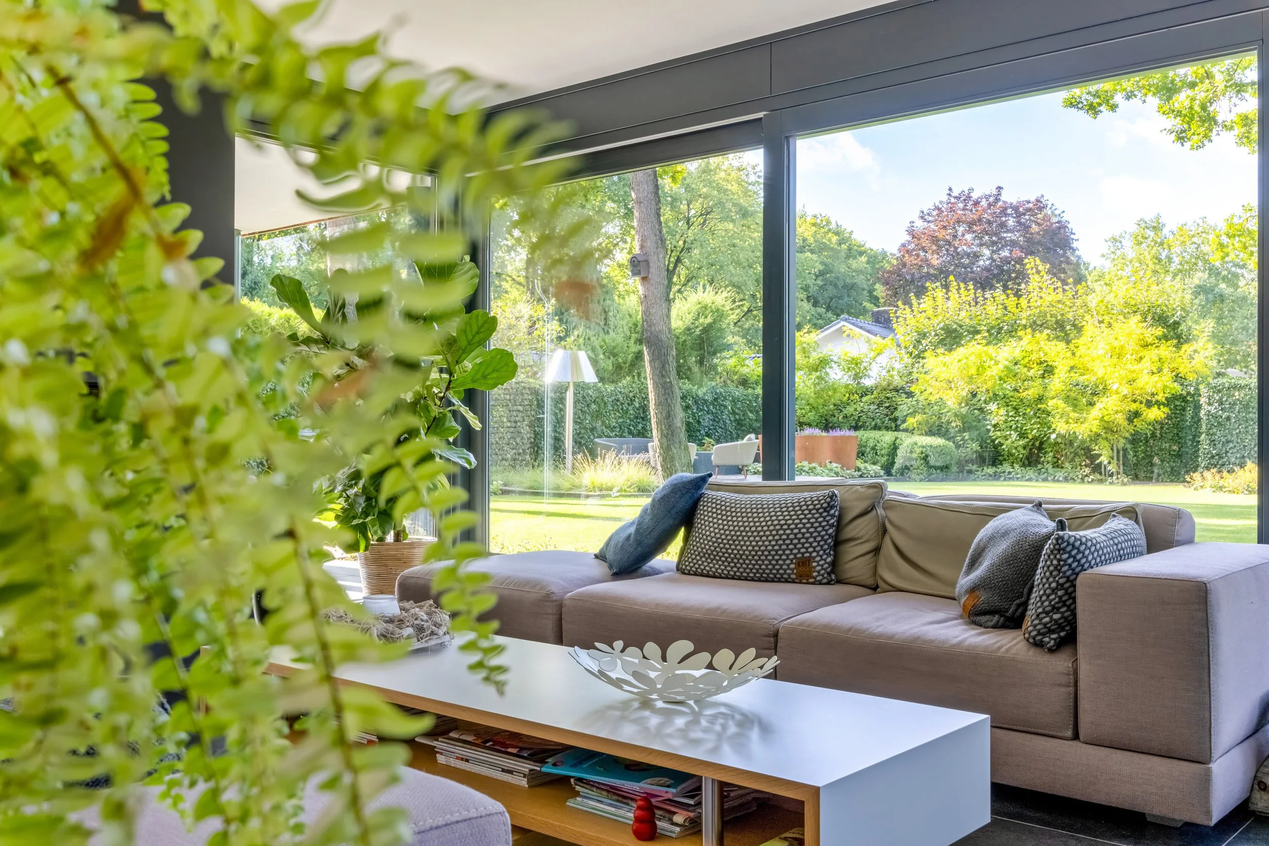 Leuk ingerichte woonkamer met grote glazen ramen, grijze bank met kussens, witte salontafel, en uitzicht op een tuin met groene planten en bomen.