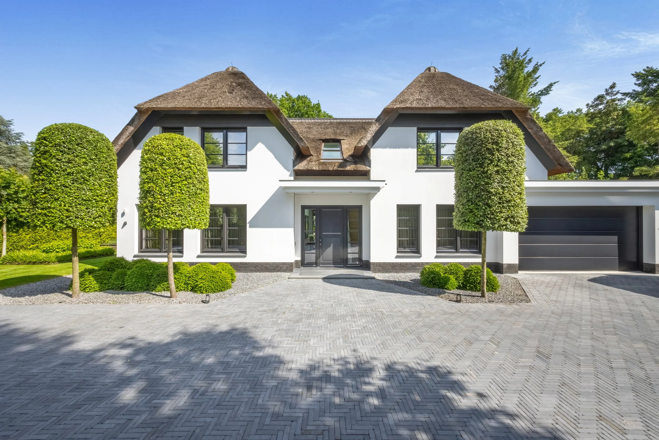 Moderne witte huis met rieten daken, twee grote getrimde bomen voor de ingang en gerestaureerde bestrating.