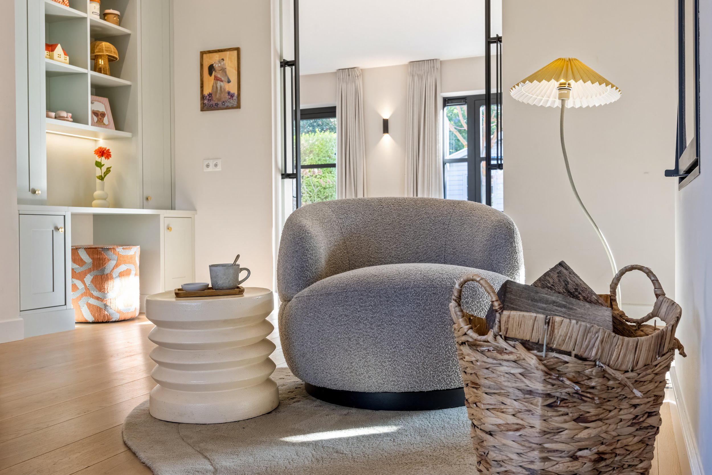 Een gezellige woonkamer met een gestoffeerd fauteuil, een ronde witte salontafel met een beker en schotel, een mand met houtblokken, een standlamp, een kast met decoratieve items, en grote ramen met witte gordijnen.