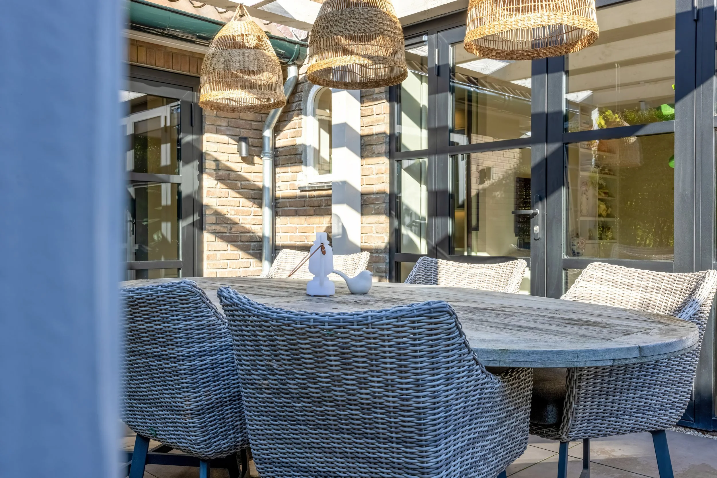 Buiten eetkamer met ronde houten tafel, zes wicker stoelen, brick muur, grote glazen deuren, hanging rieten lampen, decoratieve witte vogel en zeepaardje op tafel.