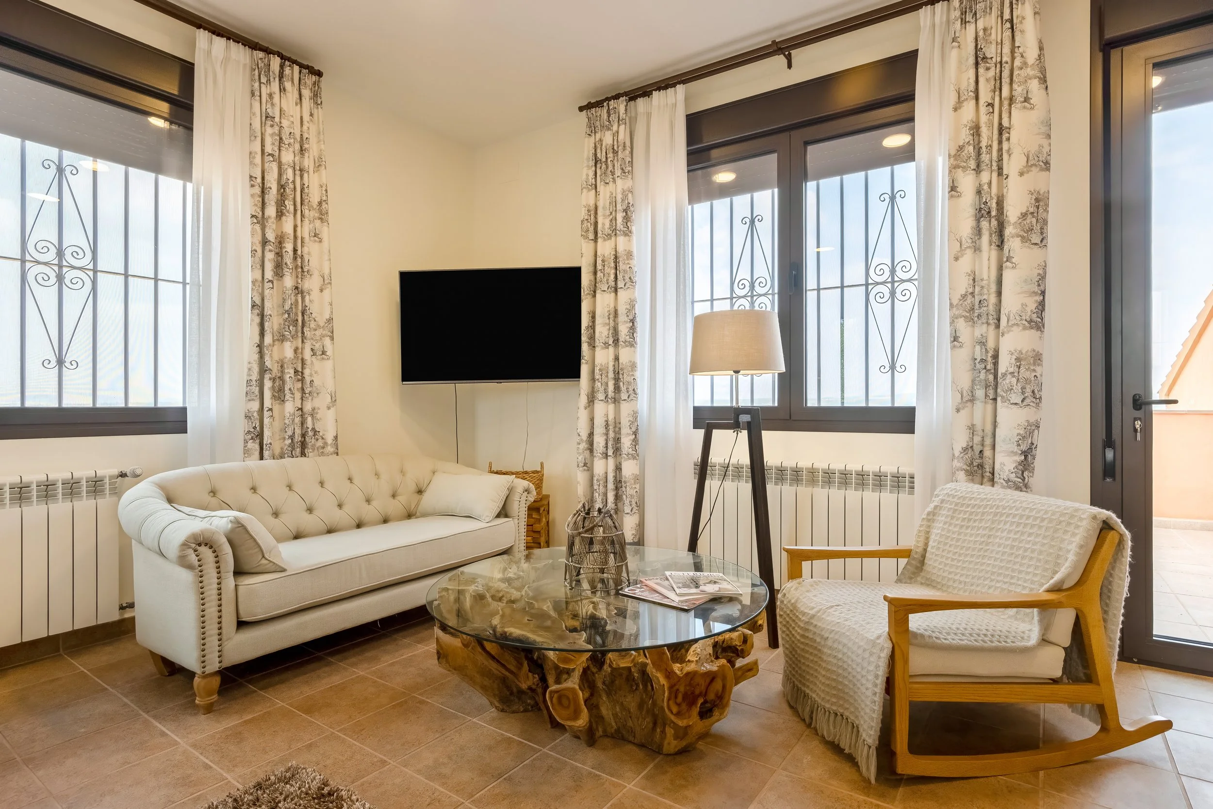 Woonkamer met witte bank, houten en stoffen armstoel, glazen tafel met houten basis, televisiescherm, lamp op standaard, ramen met witte en bloemenpatroon gordijnen, vloeren van tegels, deur naar balkon