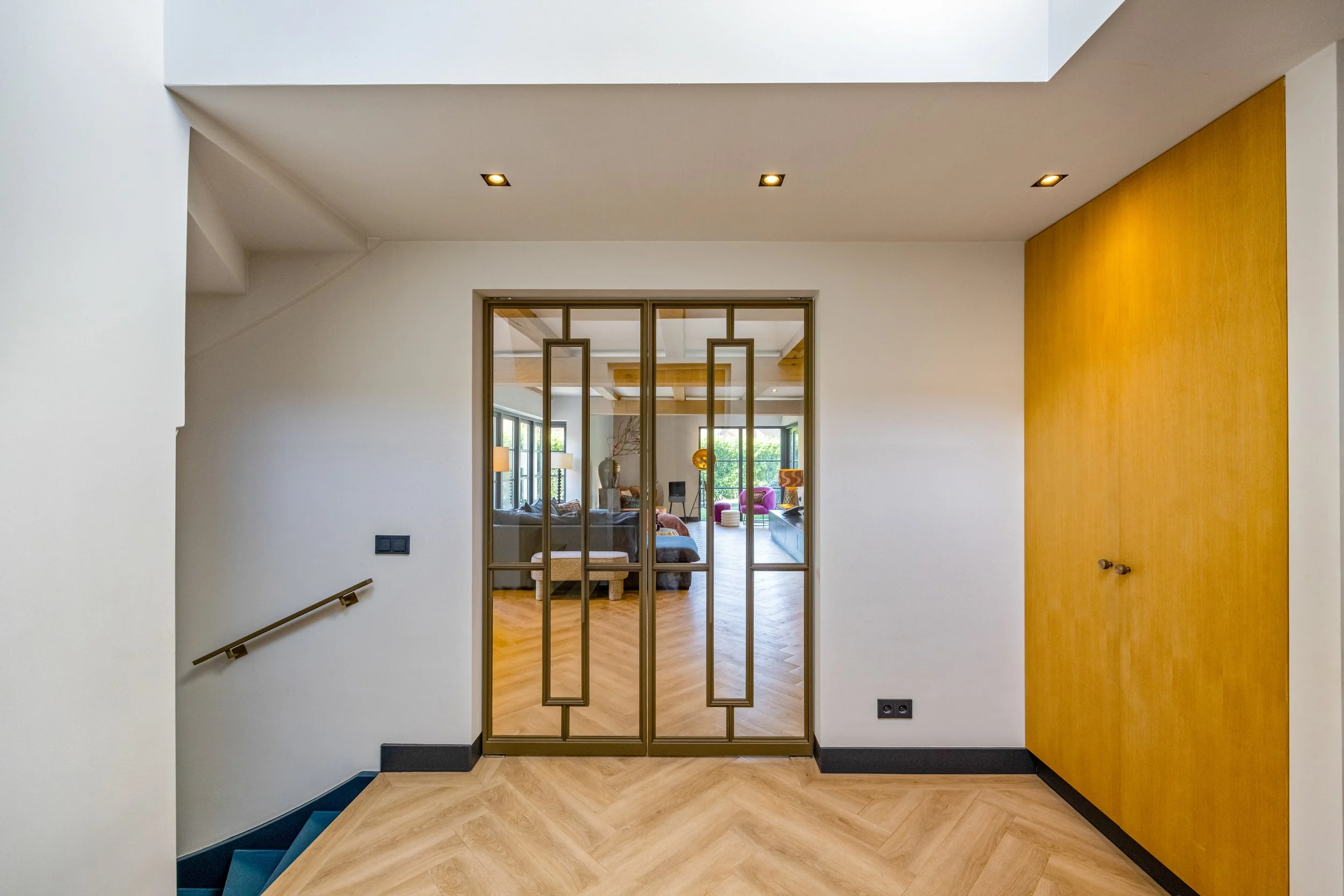 Binnenkomst in een huis met houten vloer en een glazen deur die de woonkamer scheidt, met meubels en grote ramen in de achtergrond.