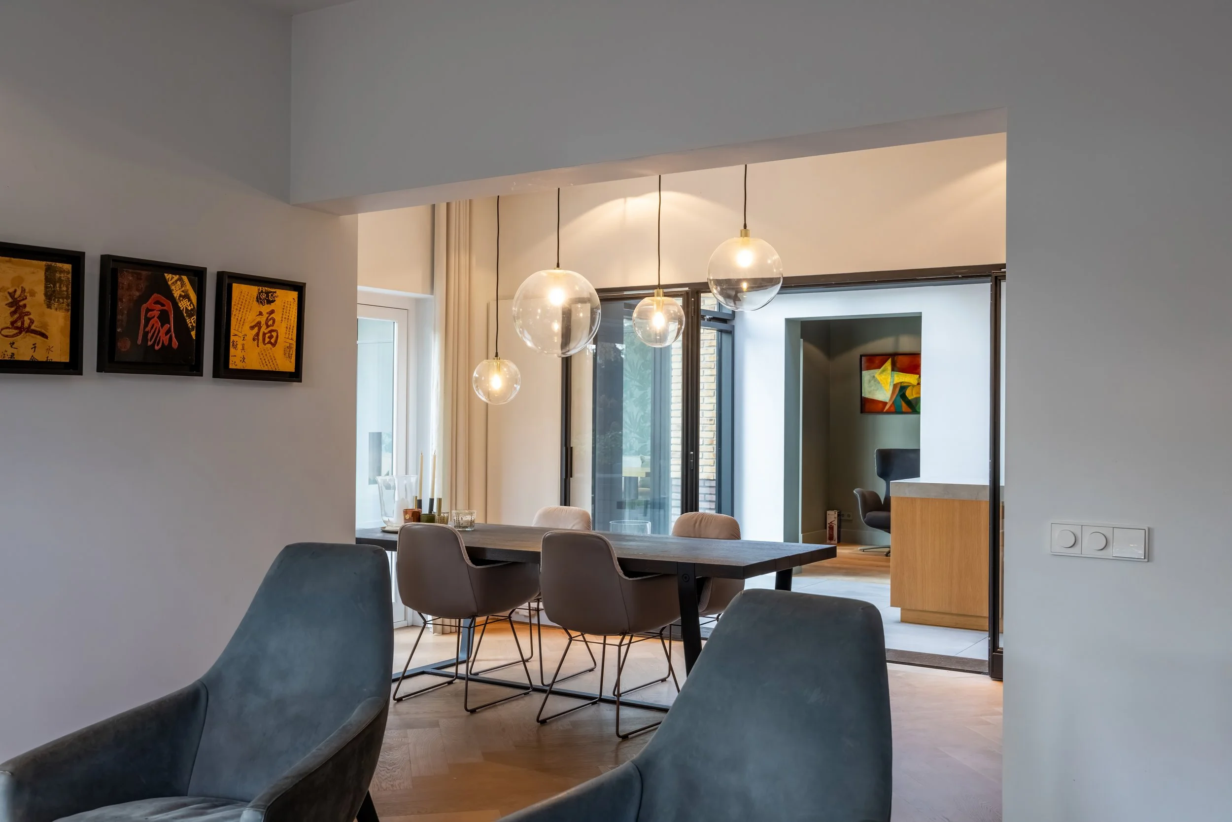 Interieur van een modern eetkamer met een houten tafel, laaghangende hanglampen, omringd door beige en blauwe stoelen, grote ramen en een deur naar een andere kamer met een kleurrijk schilderij.