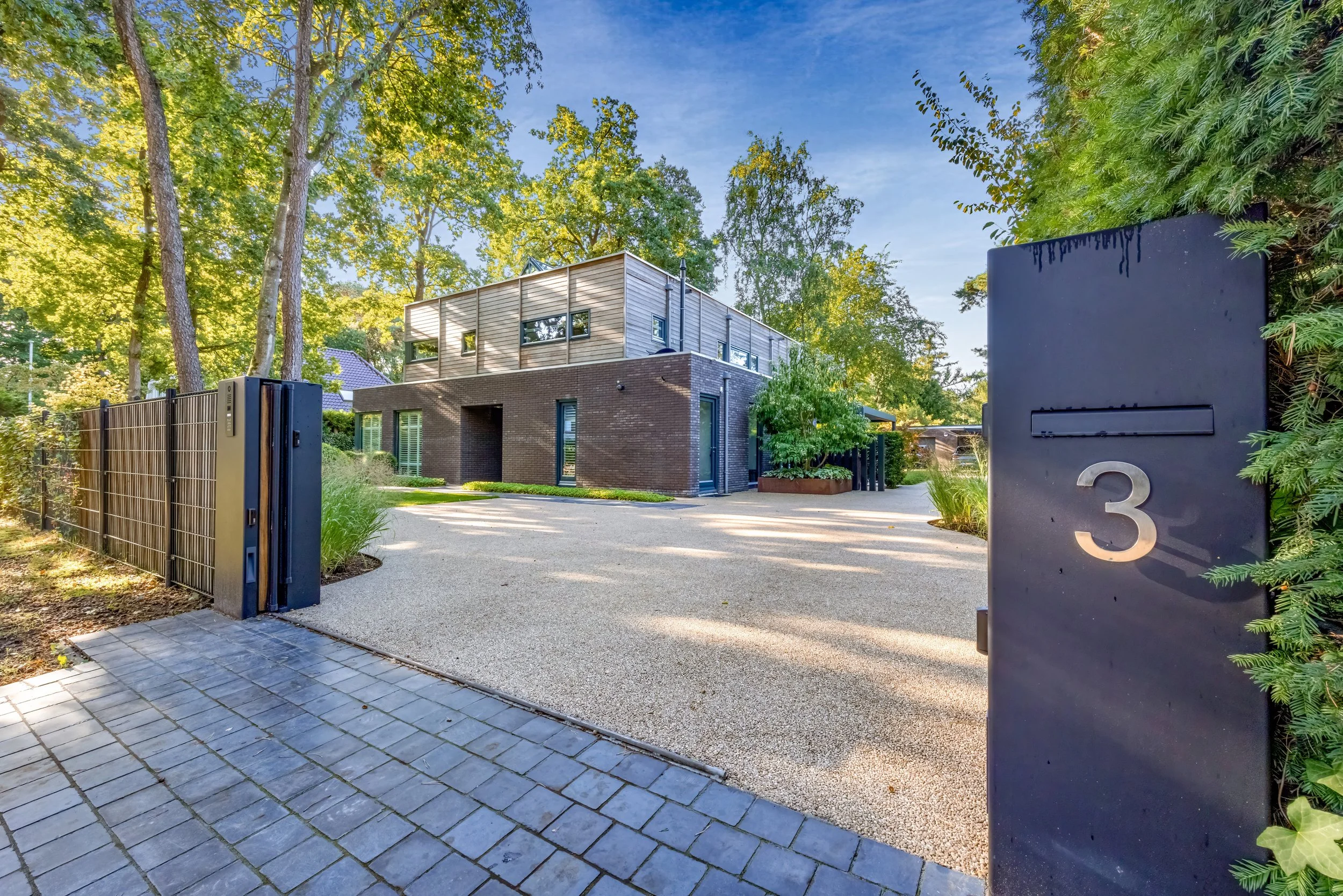 Moderne woning met een combinatie van baksteen en houten gevel, omgeven door veel groen en bomen, en een oprijlaan met een zwarte poort en een huisnummer 3.