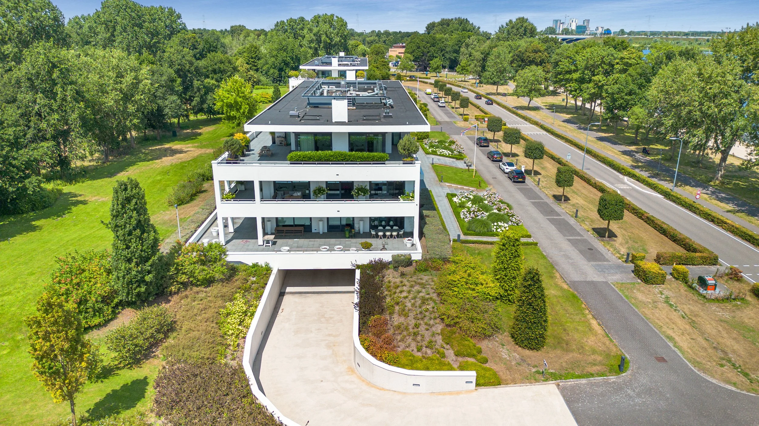Luchtfoto van een modern wit appartementencomplex met meerdere verdiepingen, omgeven door groene tuinen, parkeerplaatsen en een brede weg met bomen, in een stedelijk gebied onder een blauwe hemel.