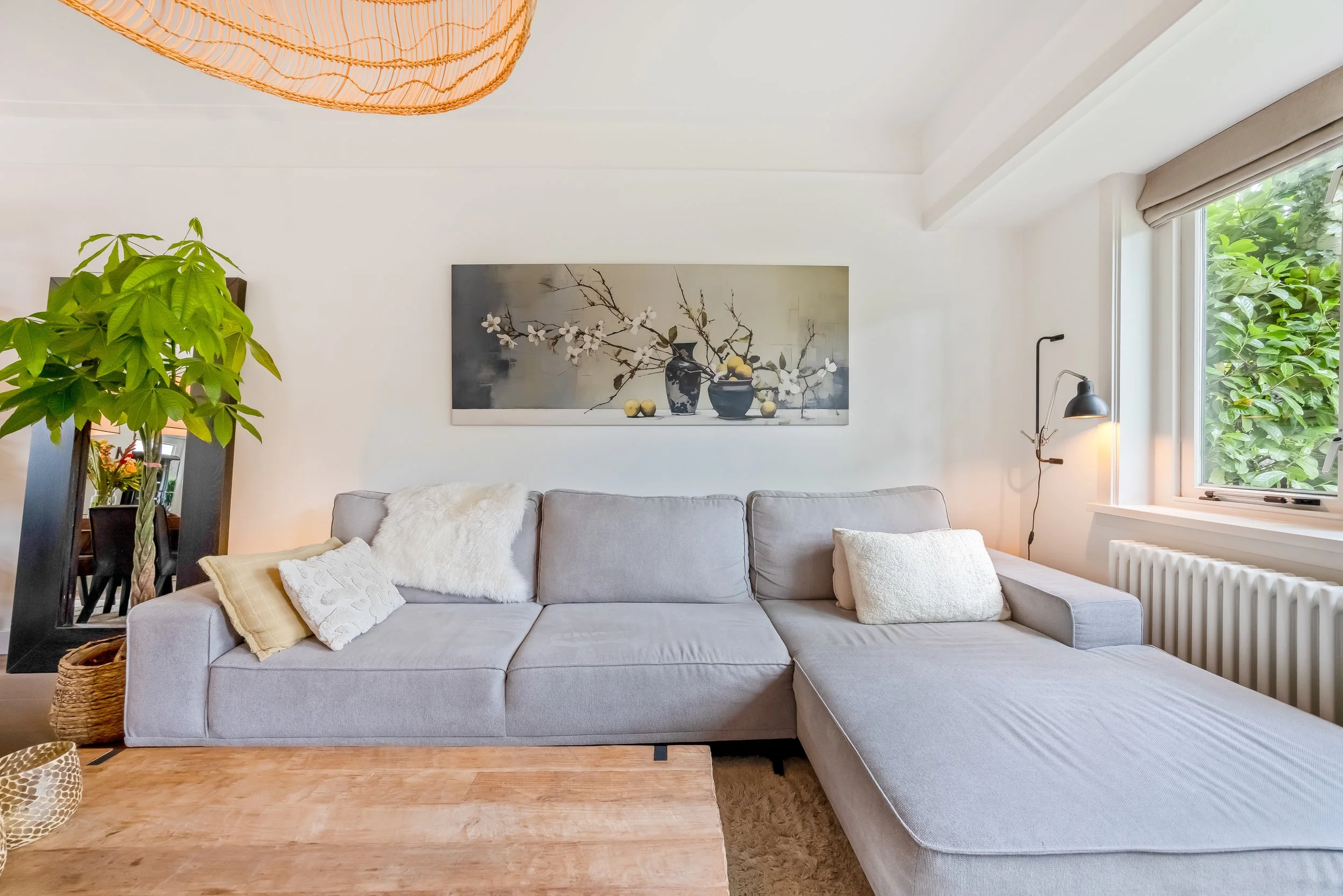 Een woonkamer met een lichte bank, decoratieve kussens, een schilderij aan de muur, een vloerlamp, een raam met groen uitzicht en een grote groene plant in een zwart frame.