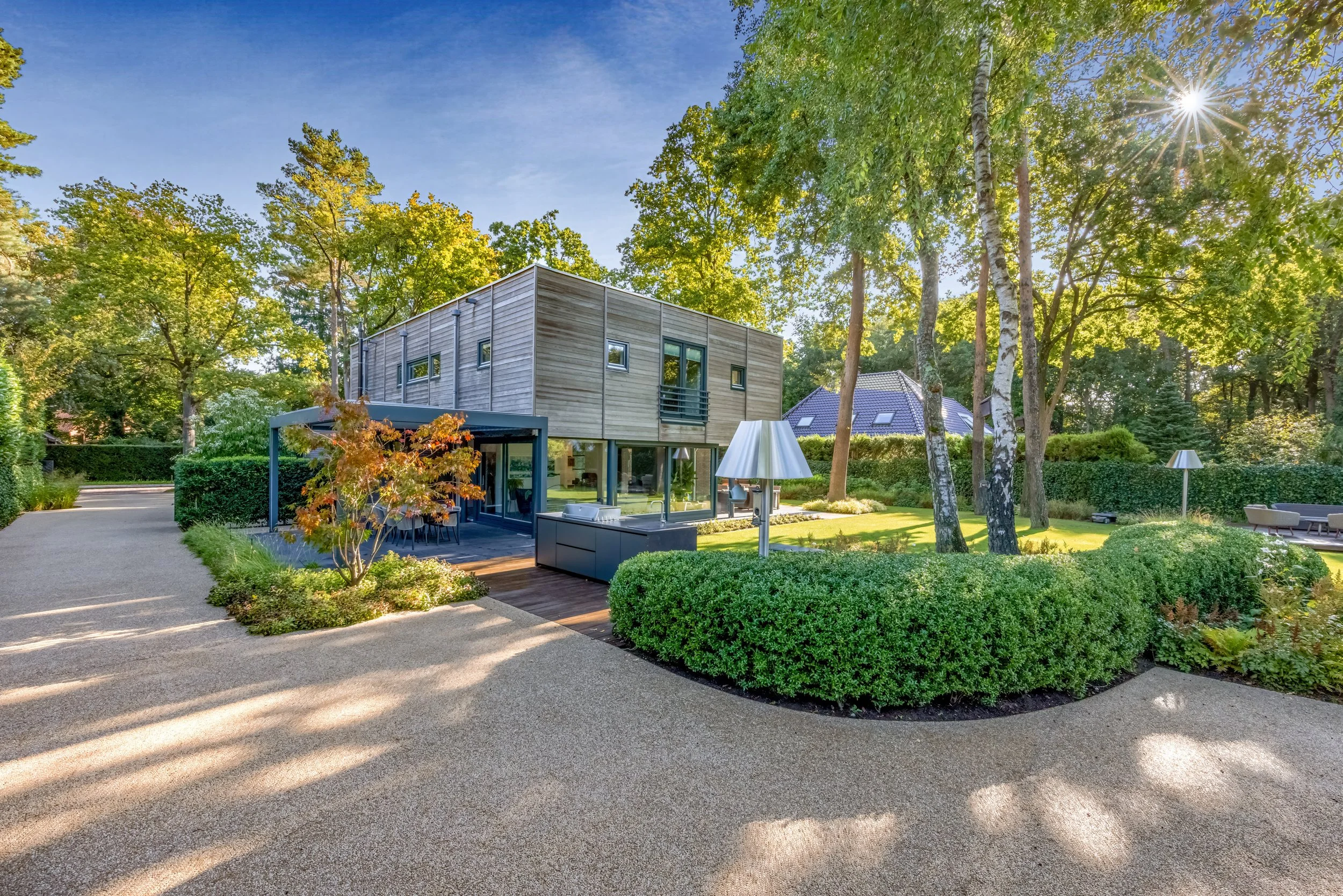 Moderne woning met houten gevel, omgeven door groene tuin en bomen, met terrasje en banken onder een grote boom, en een zonnige dag.
