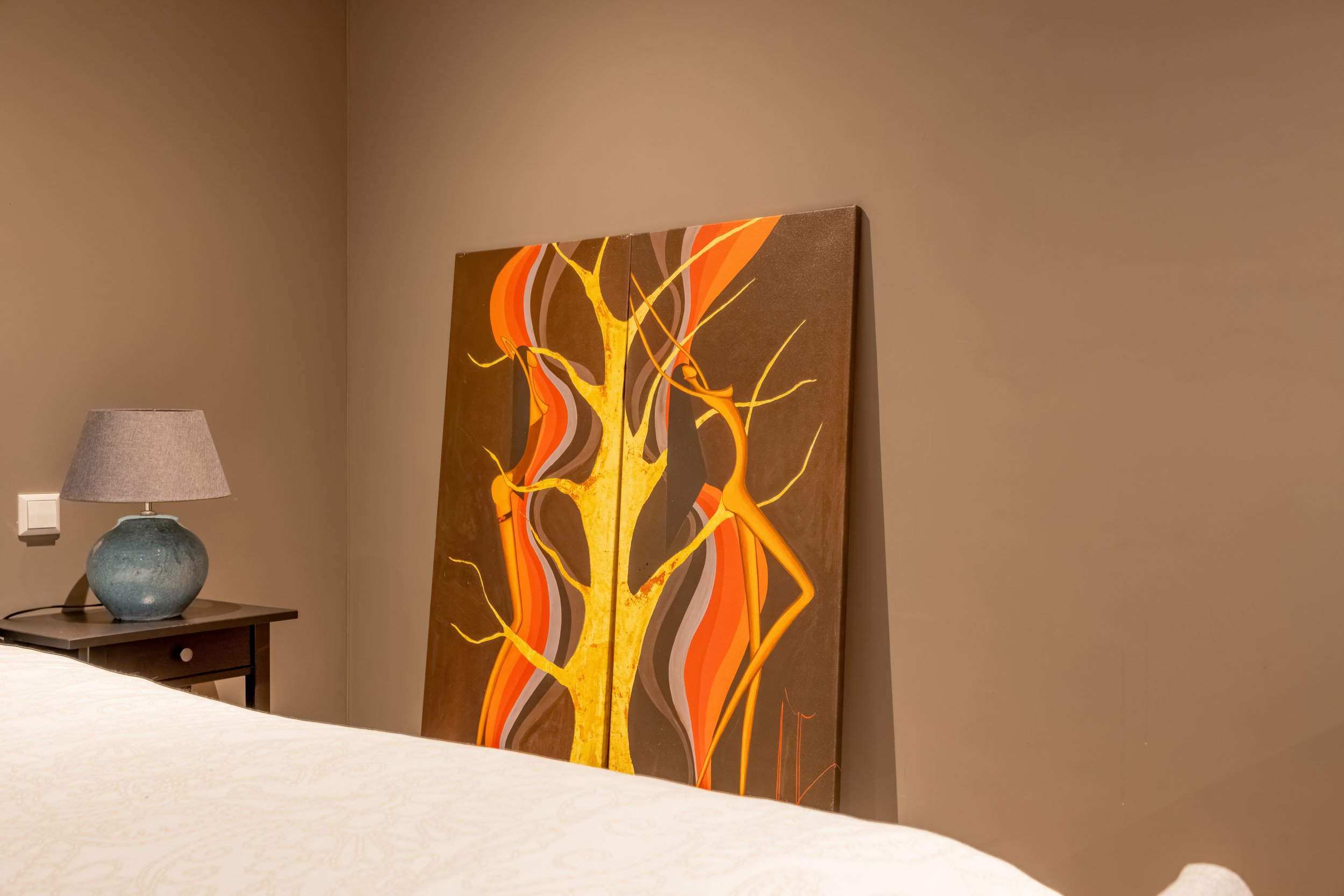 Een kamer met een beige muur, een bed met wit dekbed en een bijzettafel met een blauwe keramieken lamp. Naast de lamp staat een grote, kleurrijke schilderij met een abstract ontwerp van een gele boom en oranje en rode lijnen.