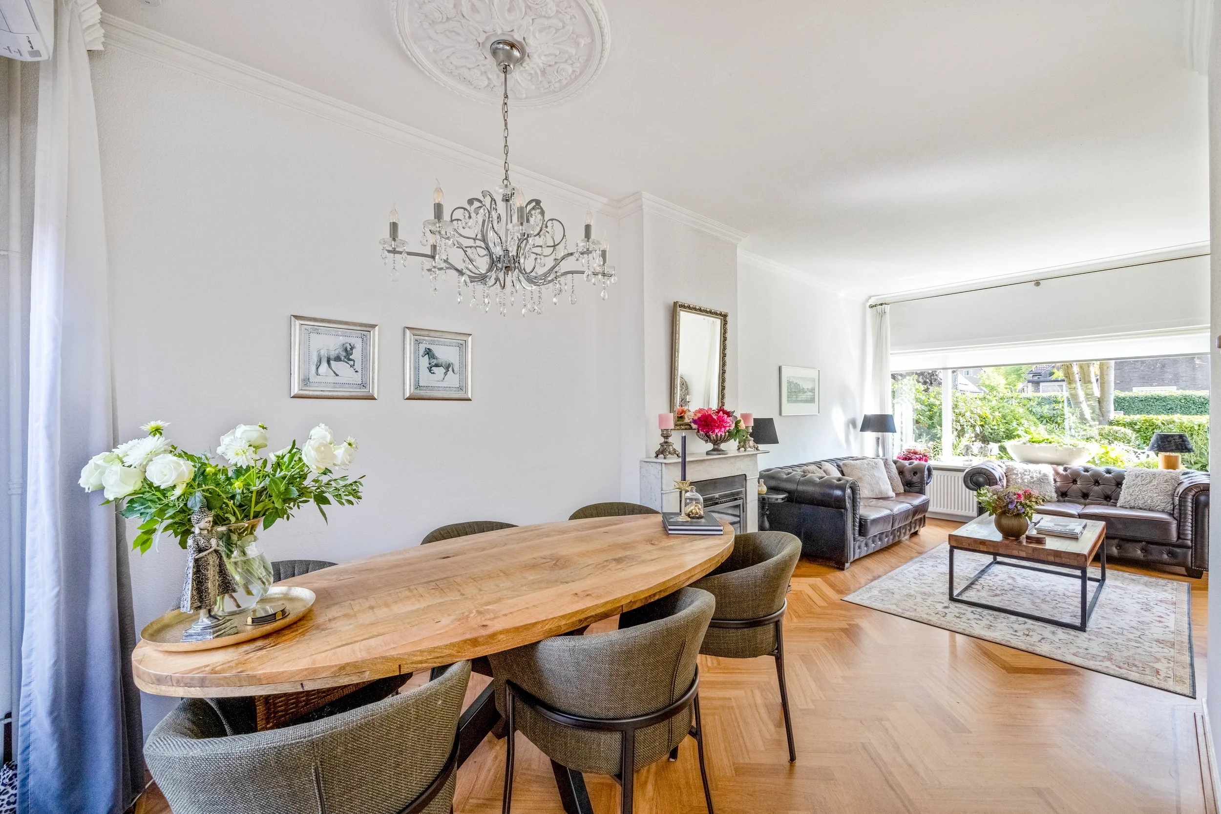 Een woonkamer en eetkamer met een houten eettafel, vijf stoelen, een sierlijk plafond met chique hanglamp, een open haard met een spiegel en bloemen, twee banken met kussens, een salontafel met bloemen, en een groot raam dat naar een tuin uitkijkt.
