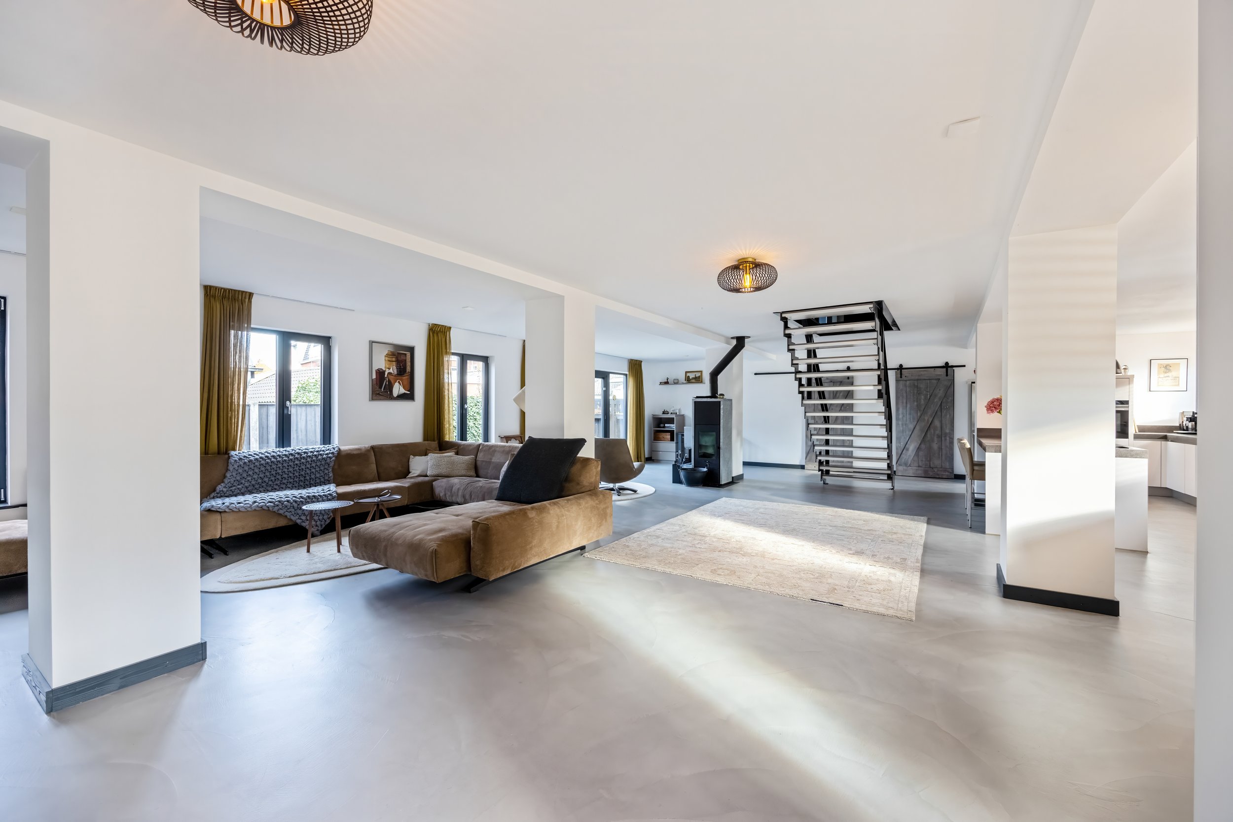 Lichte woonruimte met grote bank, meerdere ramen, open keuken uitstraling, houten trap, houtkachel, beige tapijt en modern plafondlampen.