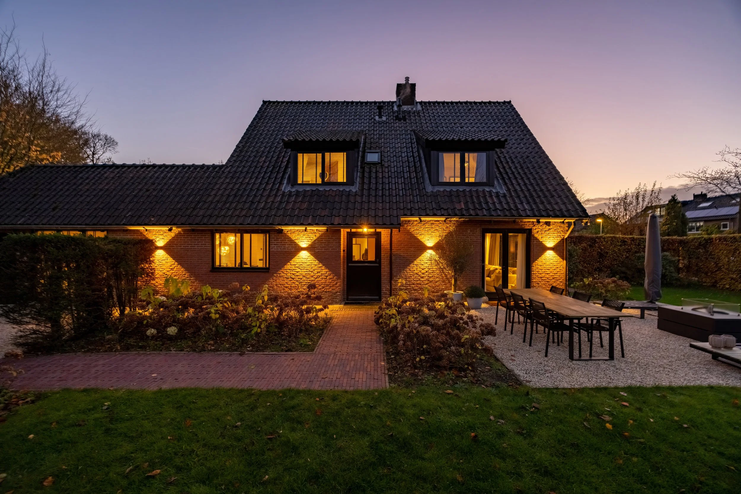 Een huis met verlichte ramen en buitenverlichting, omgeven door een tuin en een terras met tuinmeubilair, tijdens de schemering.