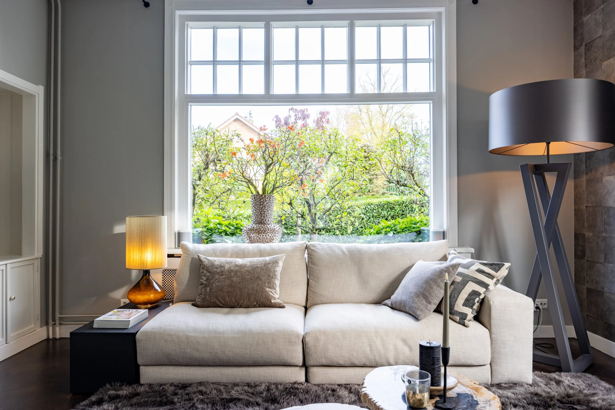 Interieur van een woonkamer met een bank, kussens, een salontafel, lampen, een grote raam met uitzicht op tuin