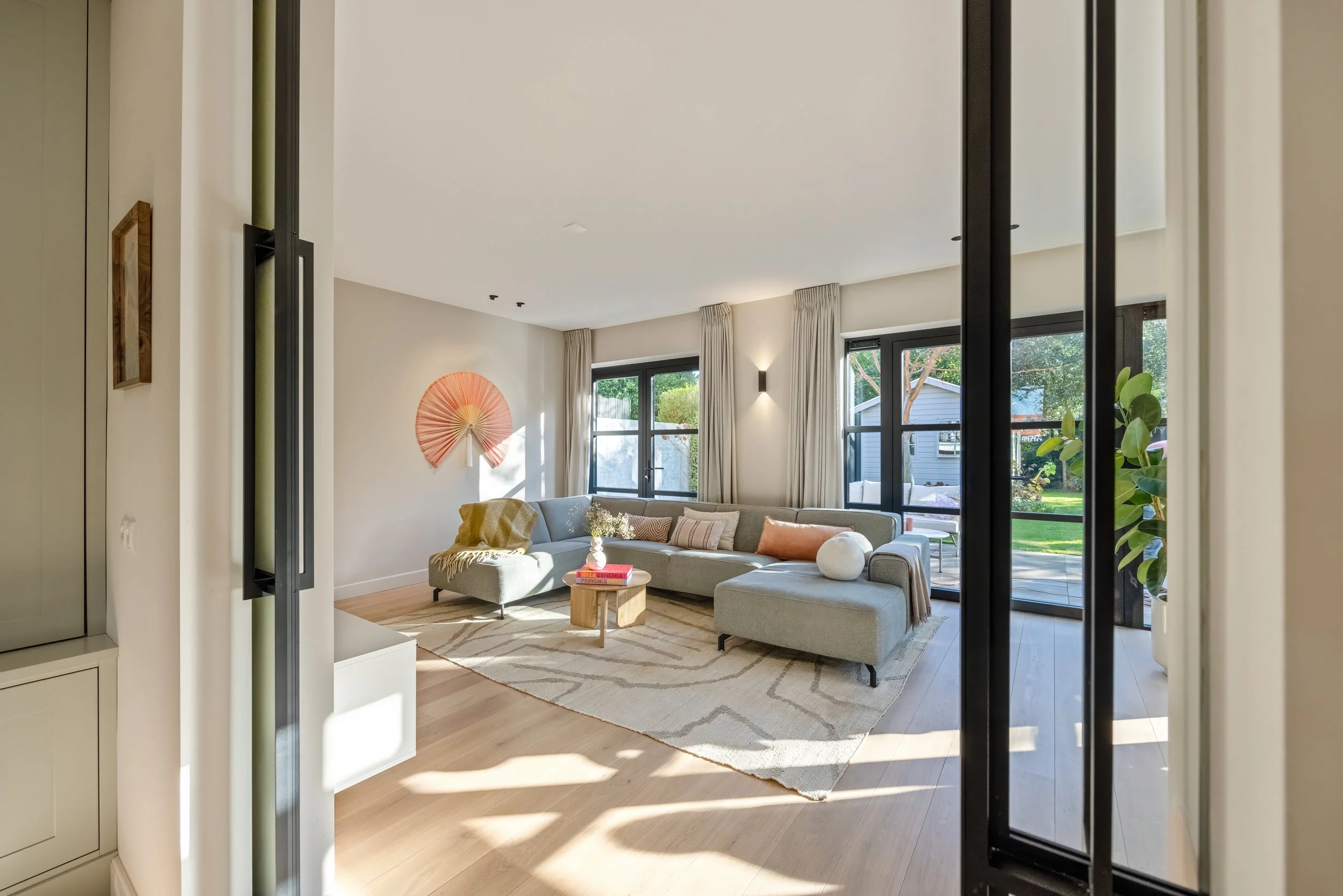 Lichte woonkamer met grote ramen, moderne bank, decoratieve kussens, salontafel, muurdecoratie, planten, uitzicht op tuin, houten vloer en oog voor design.