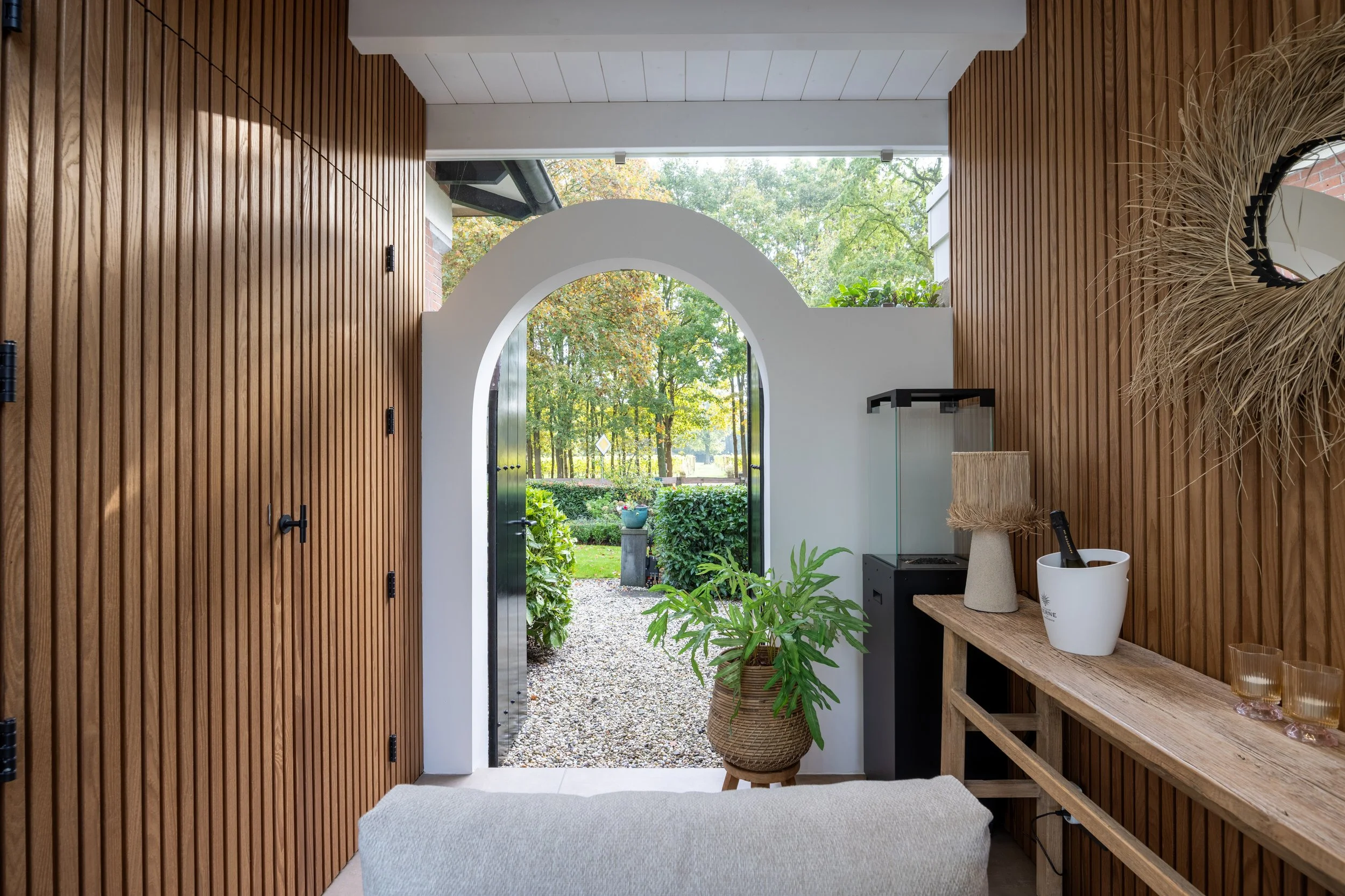 Binnenruimte met houten wandpanelen, een witte ronde arch door naar een tuin, een plantenbak met groene plant, een houten tafel met decoraties, een spiegel met een rieten frame, en een bank in de voorgrond.