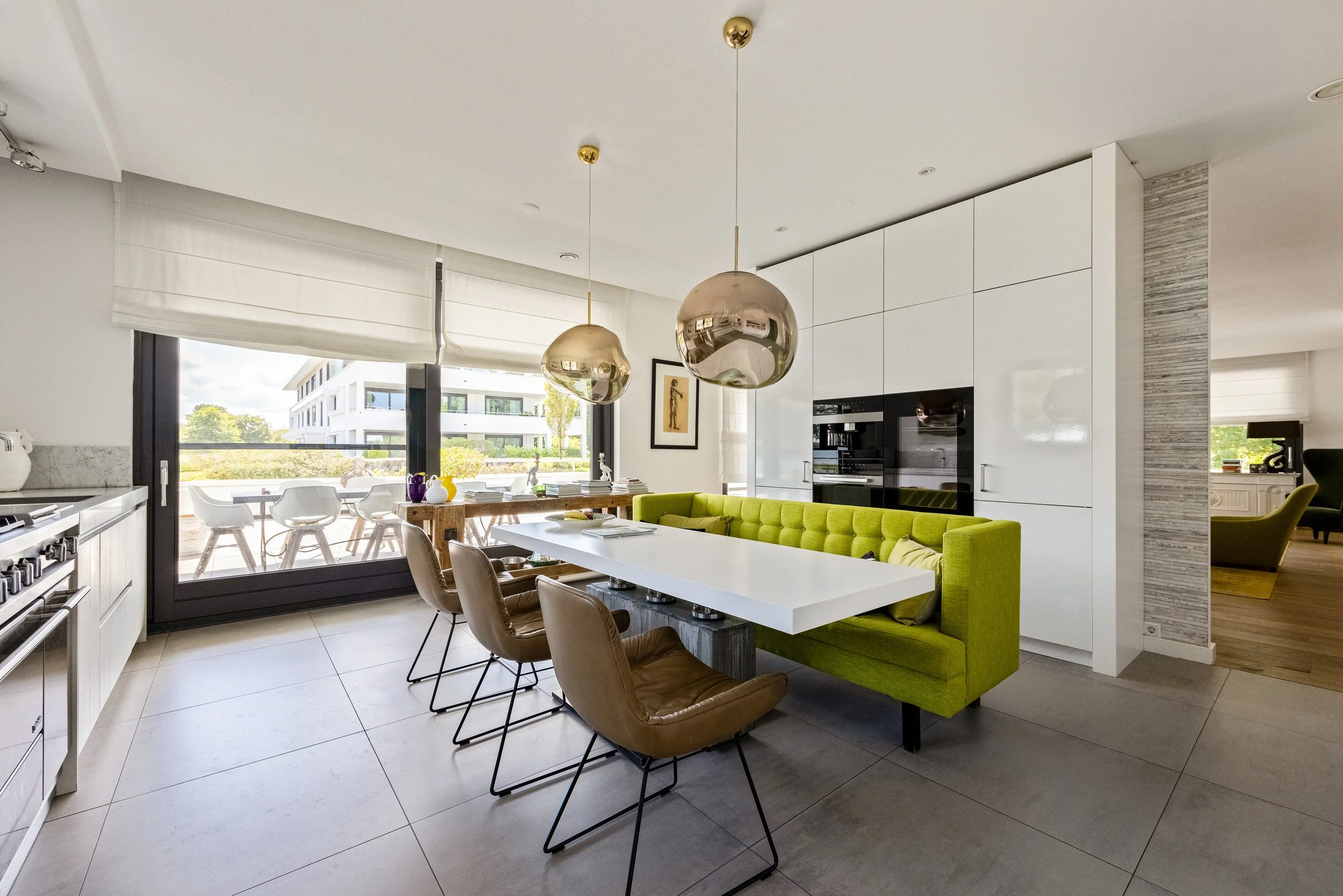 Modern keuken en eetkamer met grote witte tafel, groene bank, hangende goudkleurige lampen, grote ramen met uitzicht op balkon en tuin.