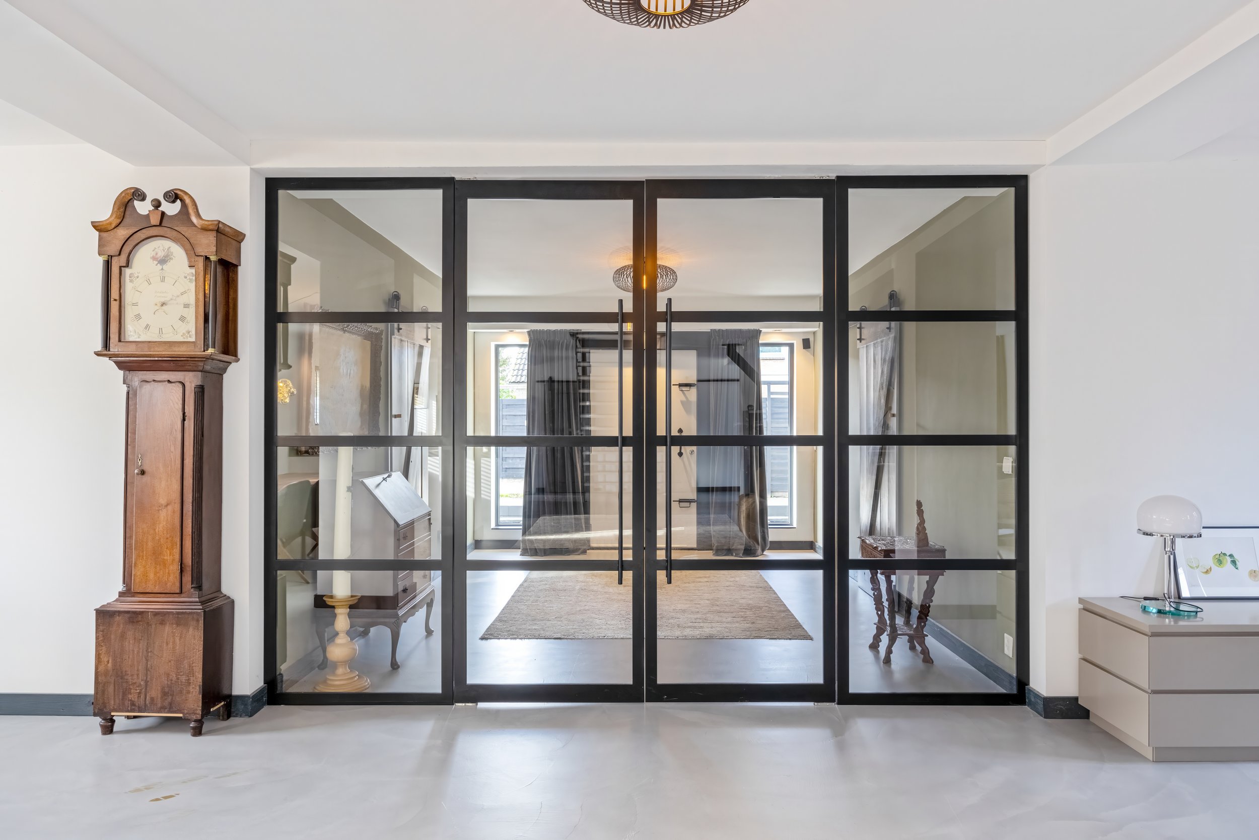 Interieur van een modern huis met een glazen deuropening, een oude staande klok links, een lichte vloer en eenvoudige meubels met een zitruimte achter de glazen afscheiding.