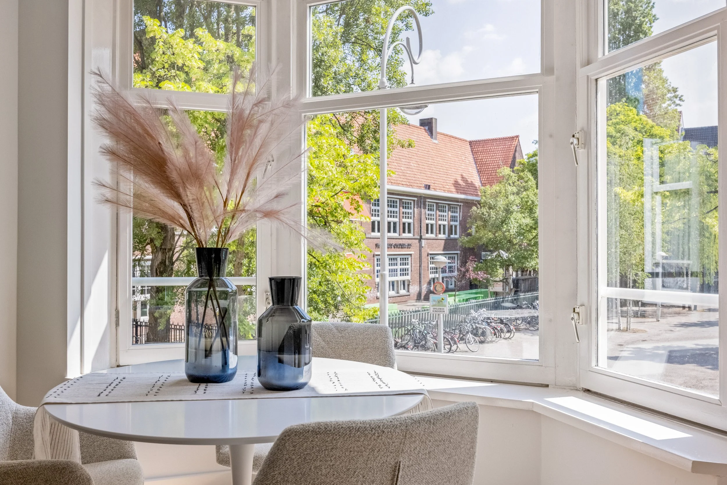 Binnenzicht op een witte tafel met twee donkere vazen en een grote tak met pluimen, naast een ramen met een uitzicht op straat en groene bomen, in een lichte kamer.