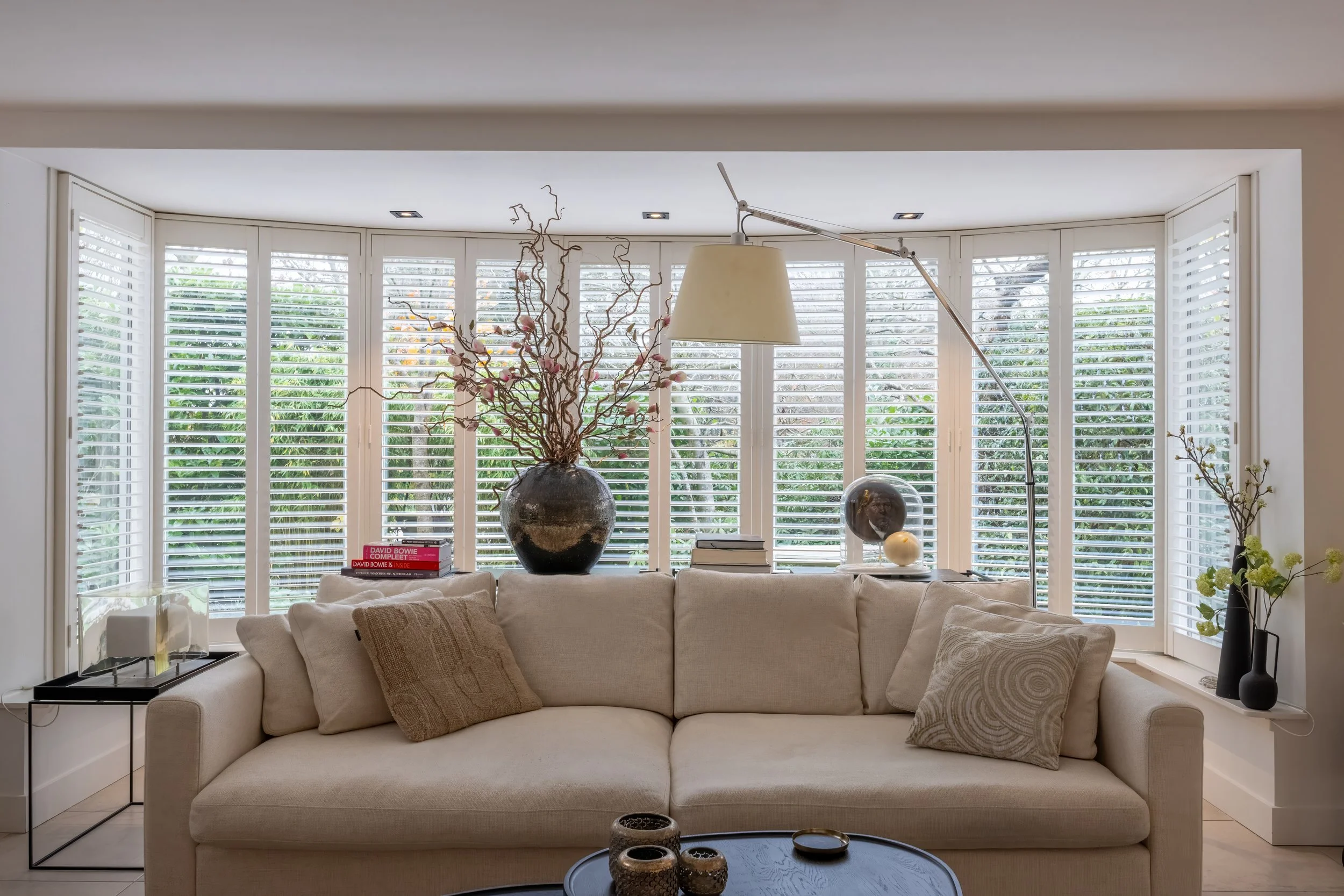 Woonkamer met beige bank, grote geschakelde ramen met witte jaloezieën, decoratieve vazen, boeken en lamp in de vensterbank.