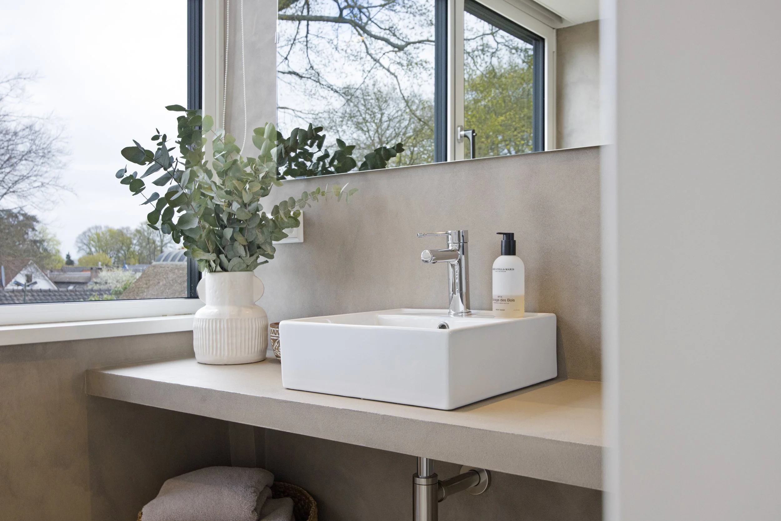 Een wit keramag wastafel op een beige betonnen aanrecht met een fles zeep en een vazen met groene planten, naast een raam met uitzicht op de buitenlucht.