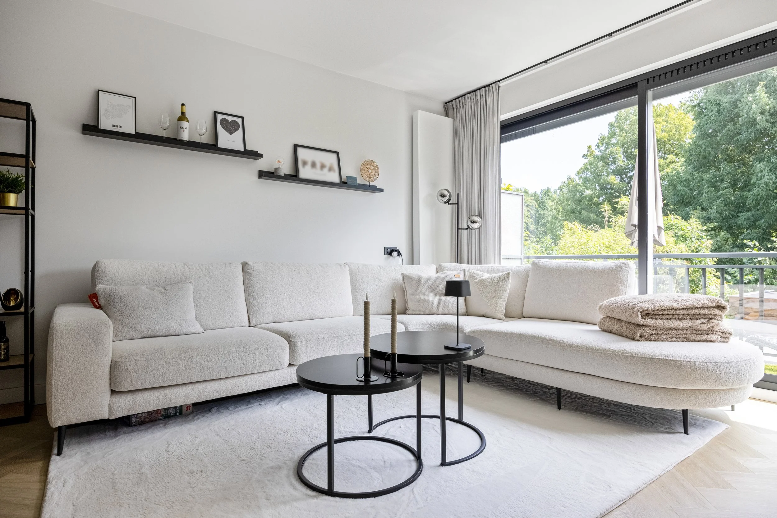 Witte L-vormige bank in een woonkamer met zwarte bijzettafels en decoraties, grote schuifpui met uitzicht op groen tuin.