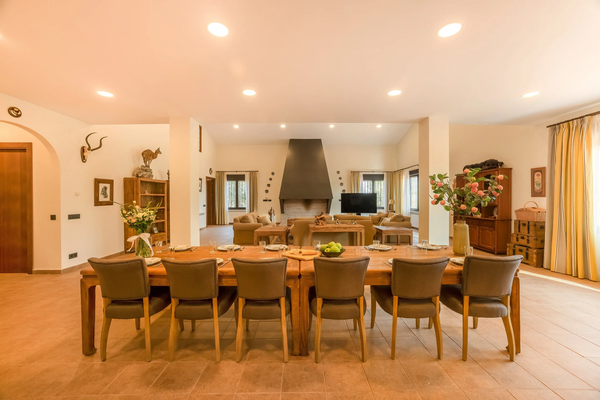 Een ruime woonkamer met een grote eettafel, acht stoelen, en een gezellige zithoek met banken en een televisie, grote ramen met gordijnen, en decoratieve stoffen en planten.