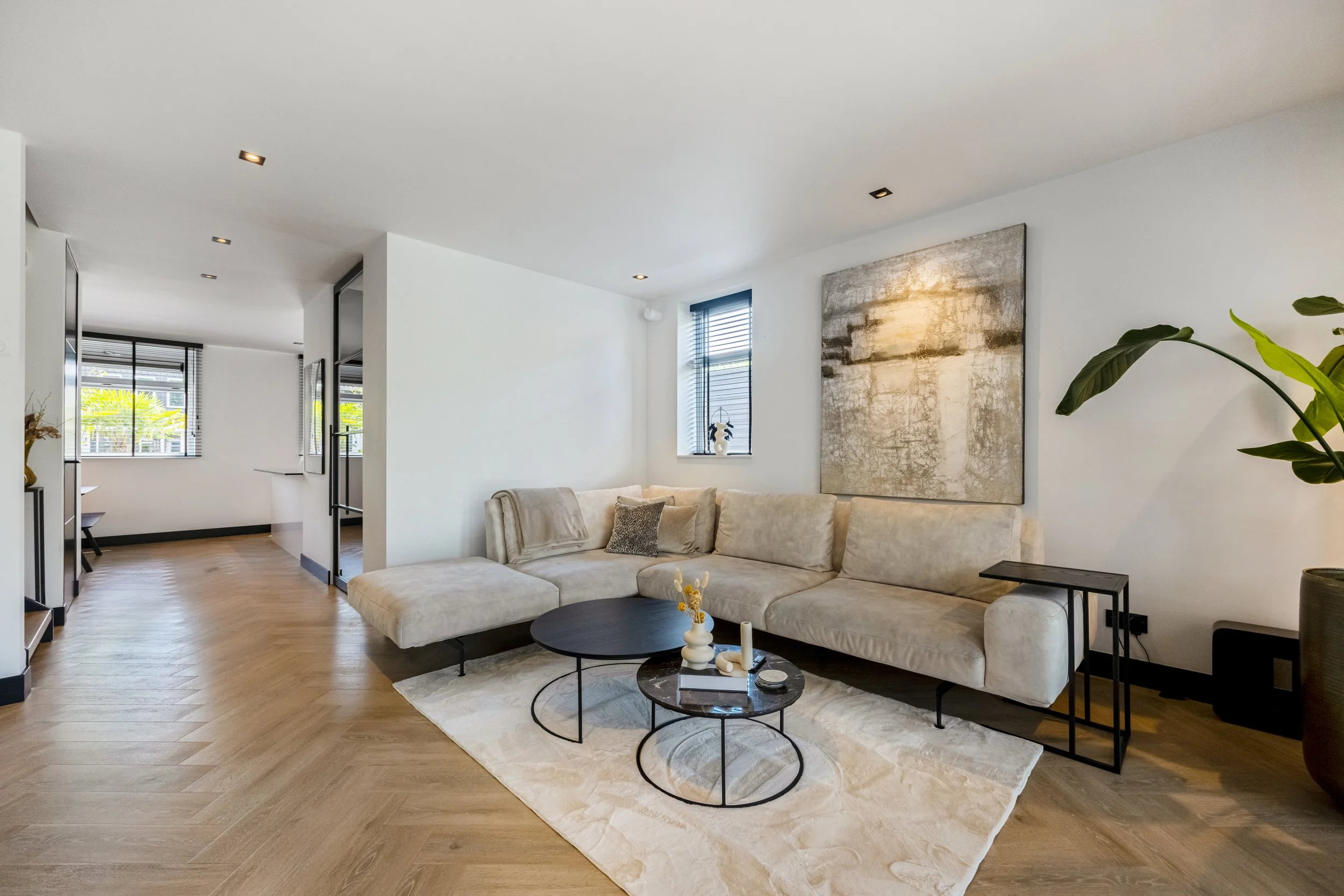 Een modern woon- en zitkamer met een ruime, lichte bank, een kunstwerk aan de muur, salontafels, een vloerkleed, en grote ramen met horren.