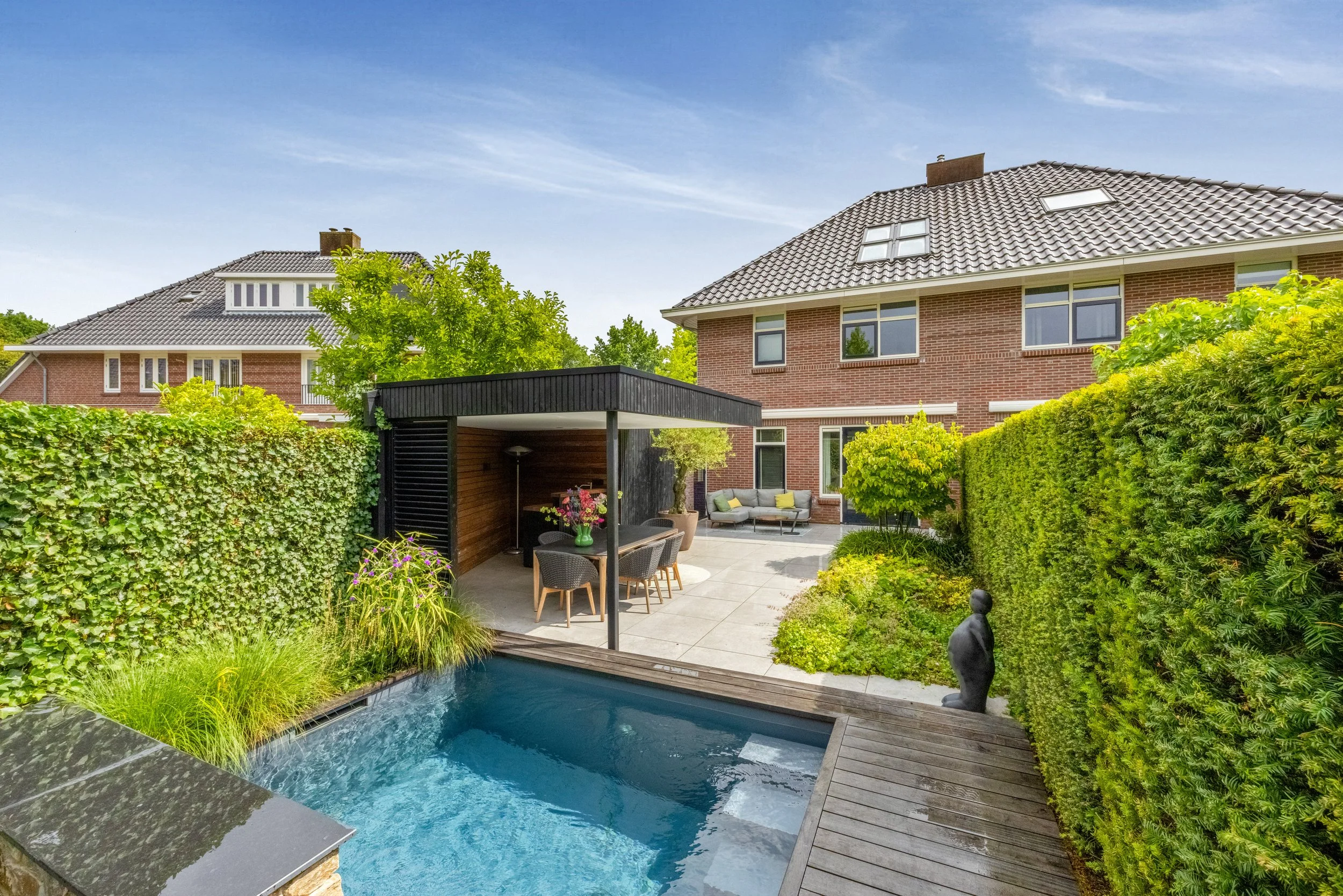 Tuin met zwembad, buitenbank, eet- en zitgedeelte, tuinbeeld, groene heggen en bomen, twee huizen met rode bakstenen muren en dakpannen, zonnige dag en blauwe lucht.