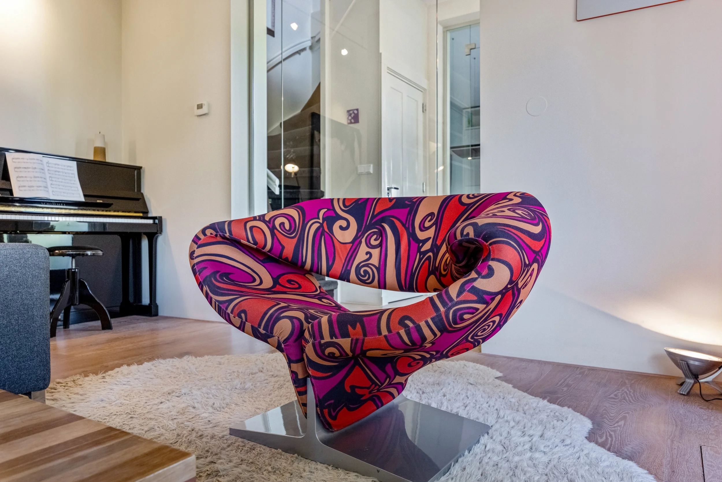 Kleurrijke, moderne loungestoel met een sierlijk design en een papegaai-achtig patroon in roze, oranje, en zwart gekleurd in een woonkamer met een witte muur, vloerbedekking en een piano op de achtergrond.