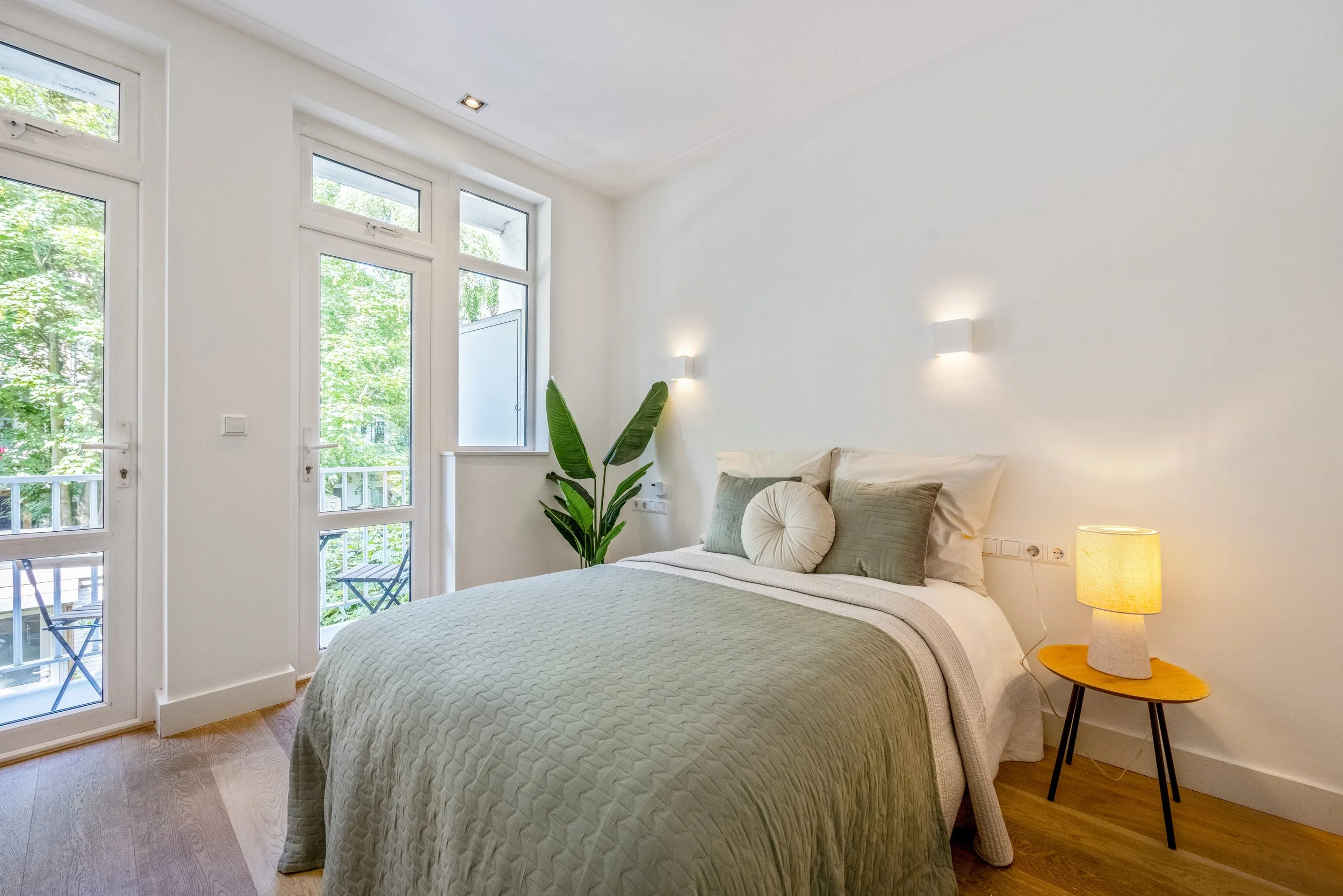 Een lichte slaapkamer met een groot bed met groen en witte beddengoed, planten en een nachtkastje met een lamp, grote ramen en een balkon met uitzicht op groene bomen.