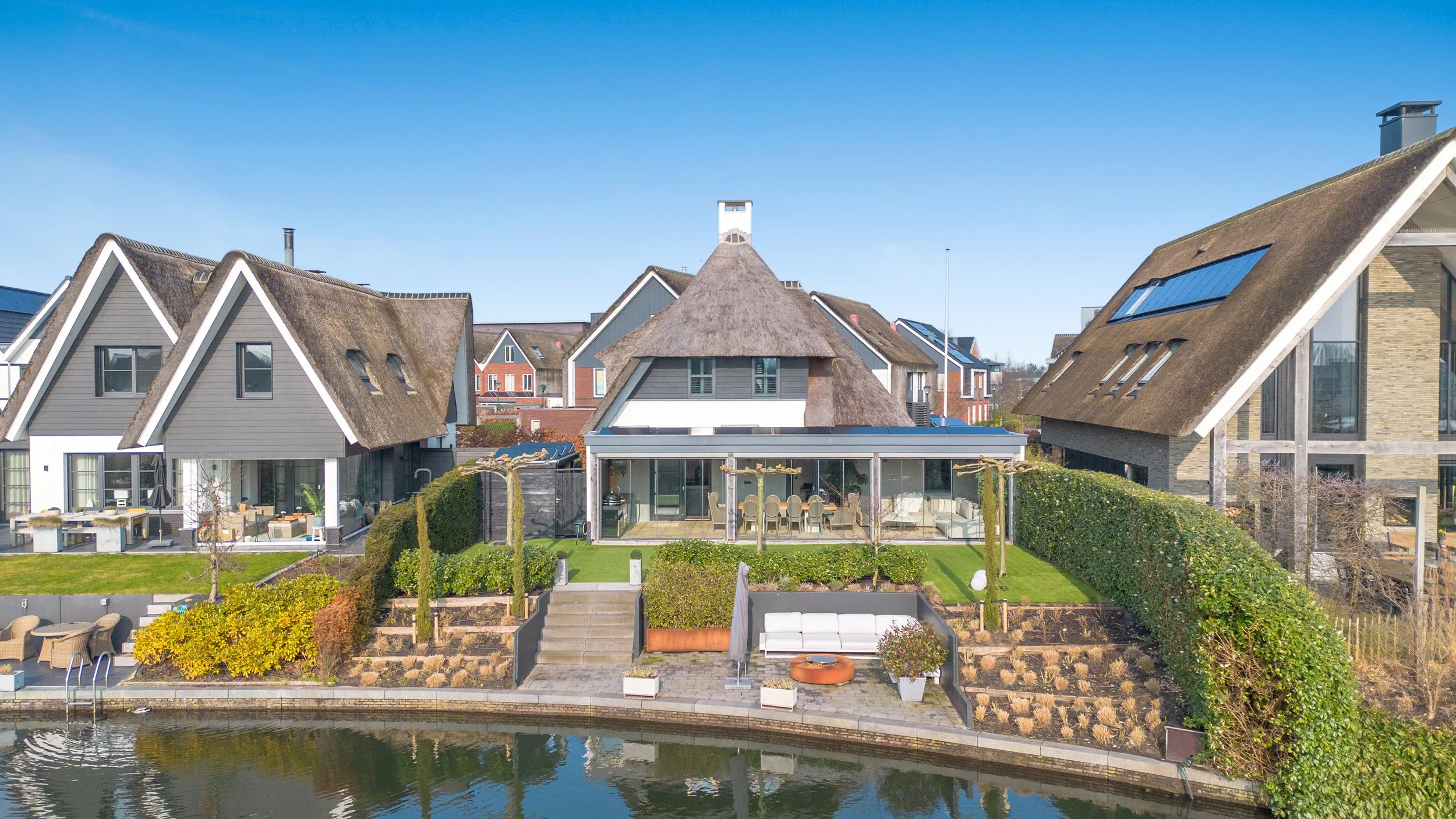 Gezicht op moderne huizen aan een waterkant met terrassen en tuin, blauwe lucht