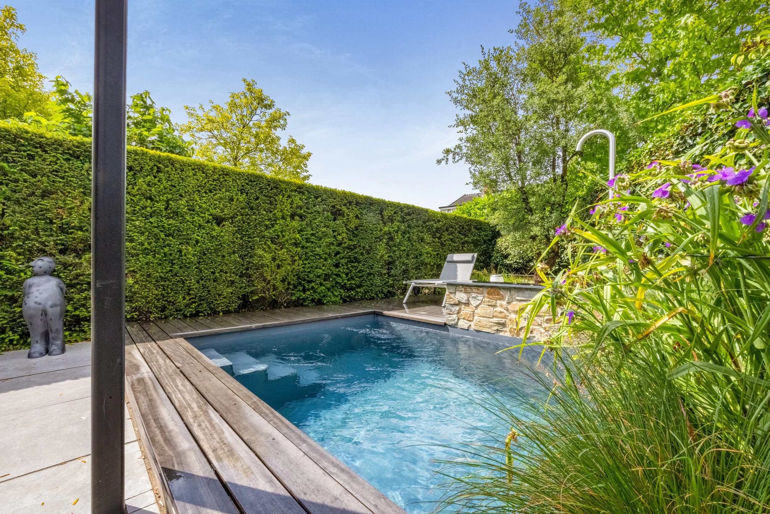 Een privézwembad in een tuin, omgeven door groen struikgewas en bomen, met een ligstoel en een fontein of waterspeelgoed op de rand.