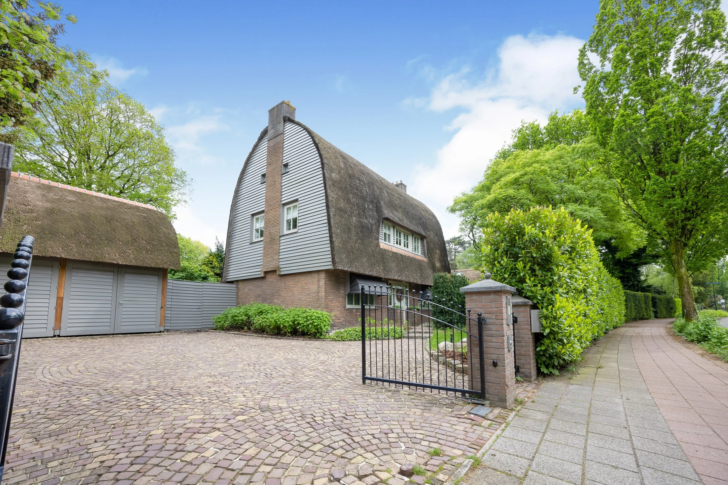Een huis met een rieten dak en een omheinde oprit, omgeven door groene bomen en een trottoir.