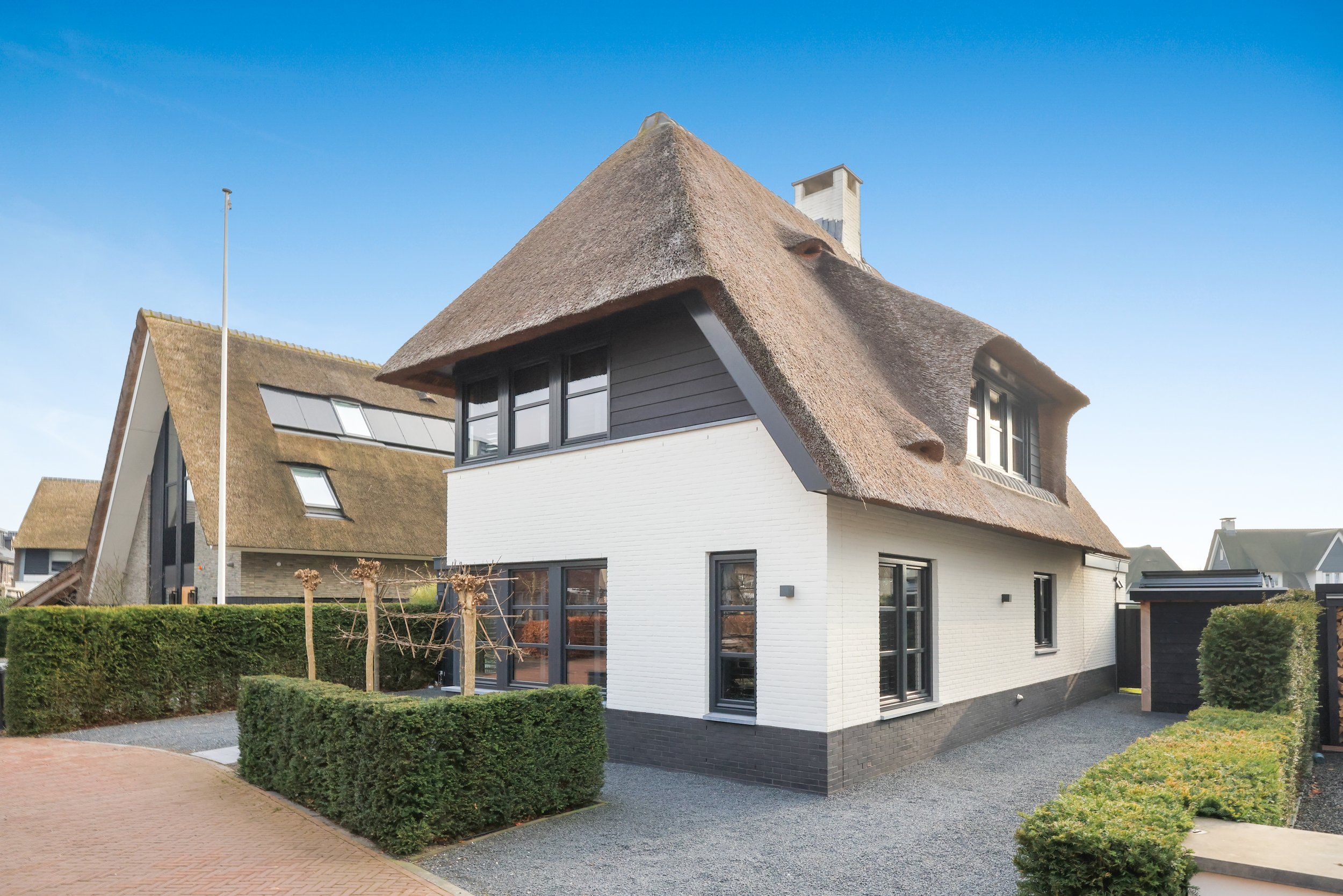 Moderne woning met een rieten dak, witte muren en grote ramen, omgeven door een tuin met heggen en een oprit, onder een heldere blauwe hemel.