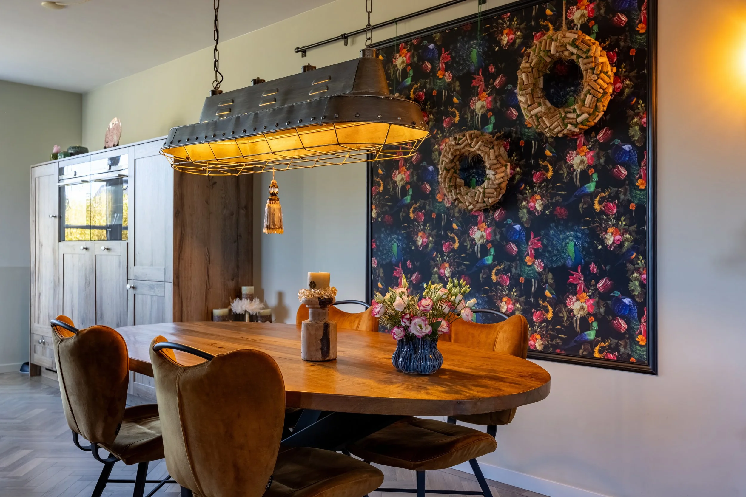 Eettafel met zeven stoelen, bloemstuk in vaas, kaarsen en decoratie, hanglamp boven tafel, muur met bloemenprint en wenskaarten, kast in de achtergrond.