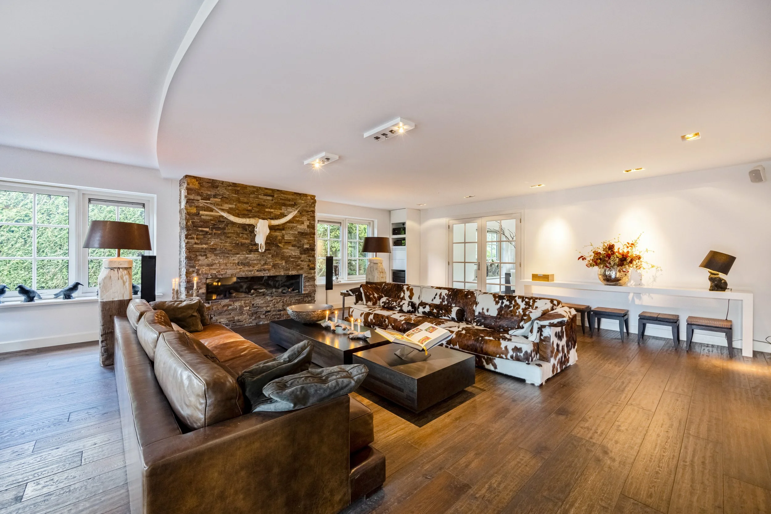Een woonkamer met een bankstel met dierenhuidpatroon, een andere leren bank, een centrale salontafel met kaarsen, een stenen haard met een gewei en een koeienschedel, en grote ramen en deuren die naar buiten leiden.
