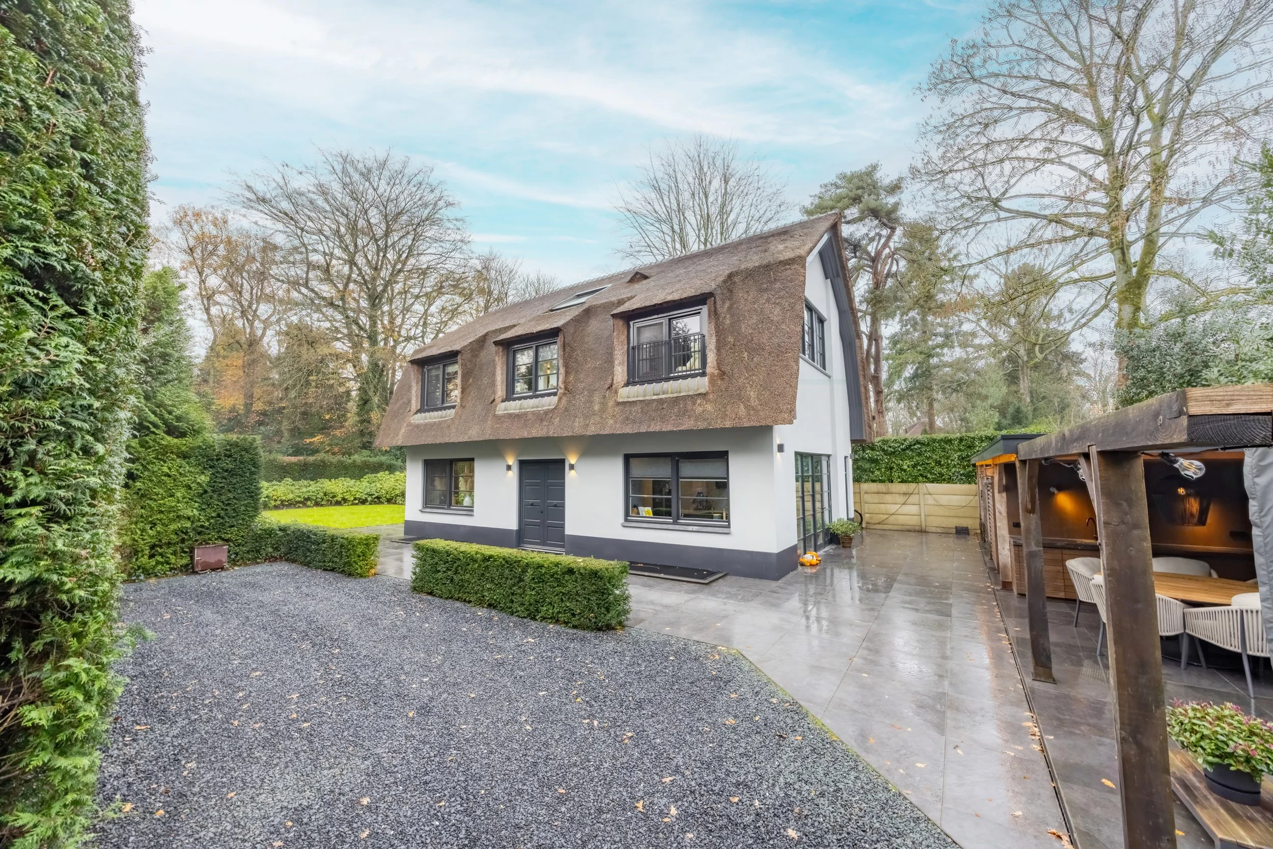 Moderne huis met rieten dak en grote tuin, gras, bomen, terras met tuinmeubilair, omringd door struiken en houten schutting.