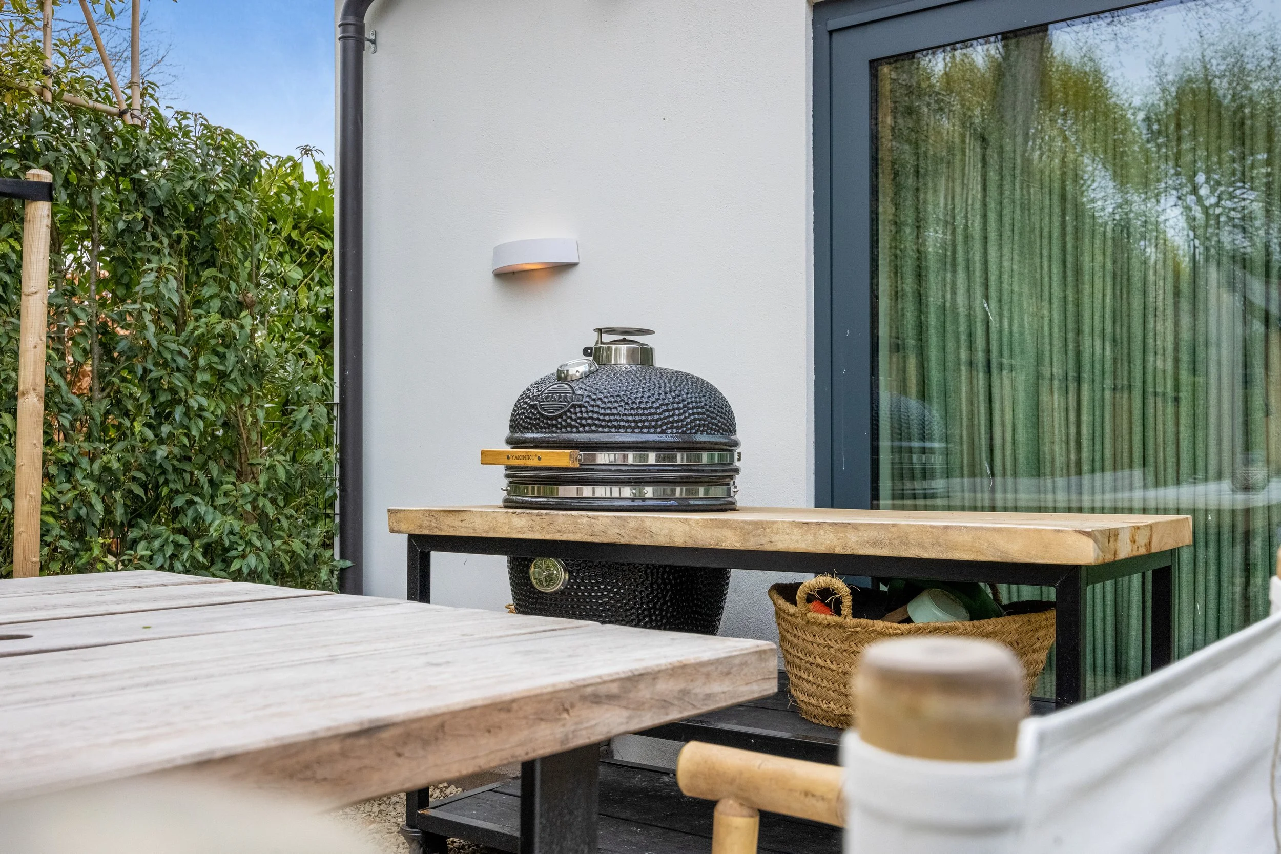 Buitenruimte met een barbecue op een houten tafel, een basket met spullen, en een schuifpui met groene gordijnen.