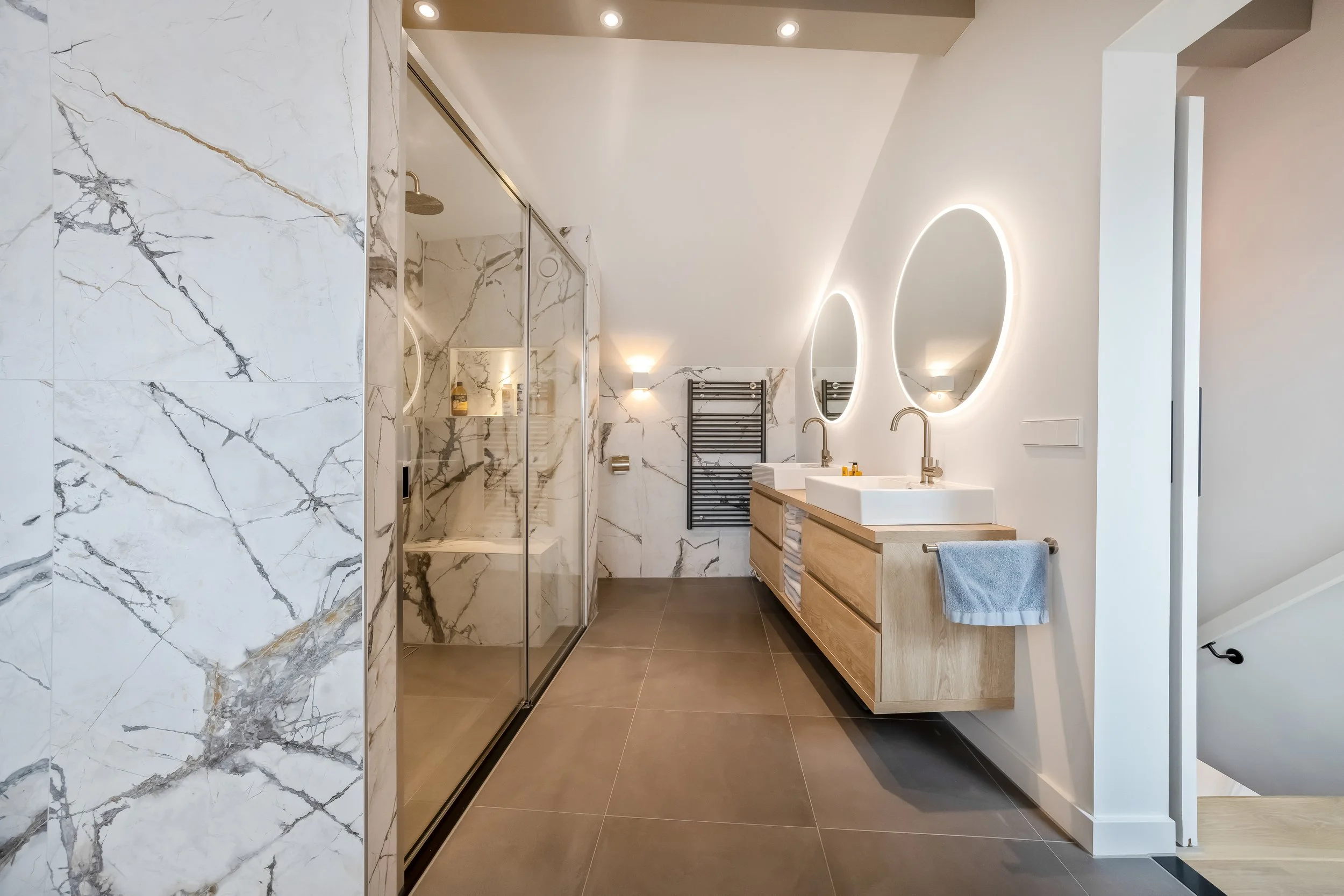 Modern badkamer met dubbele wastafel, rond verlichte spiegels, inloopdouche met glazen deur, muurpanelen van wit marmer met grijze aders, en vloer in donkere tegels.